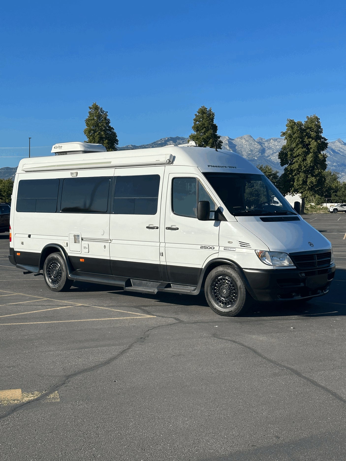 White Pleasure-Way Sprinter RV rental, parked in a lot with mountains and trees visible under a clear blue sky.