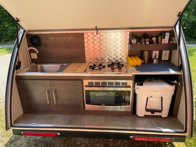 RV rental: Compact outdoor kitchen with a sink, stove, microwave, & cooler for versatile camping meal prep.