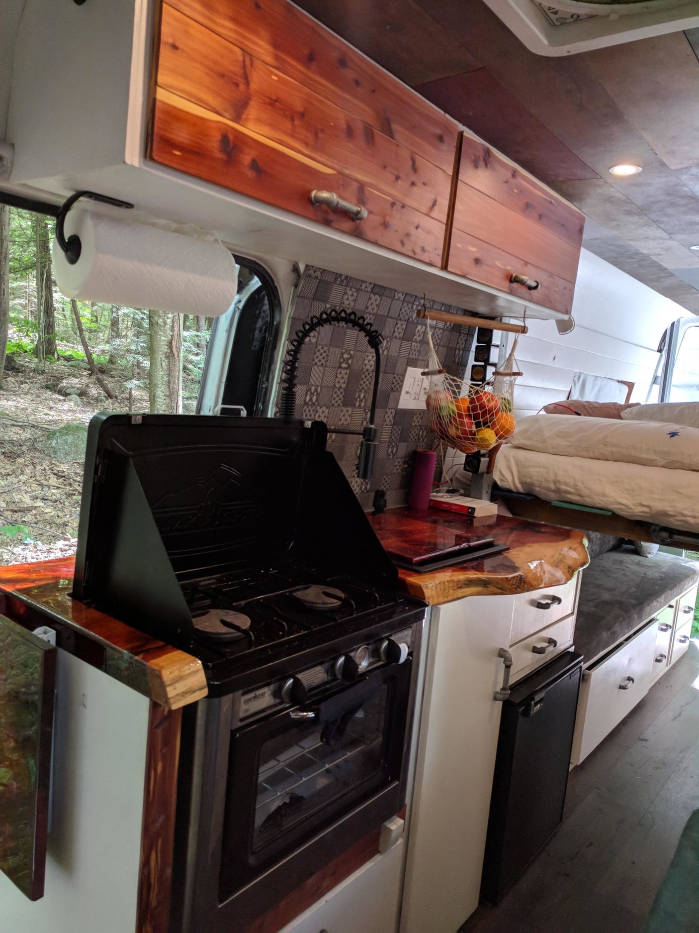 Inside a custom RV rental, showcasing a functional kitchen with stove, oven, sink, and ample storage, plus forest views.