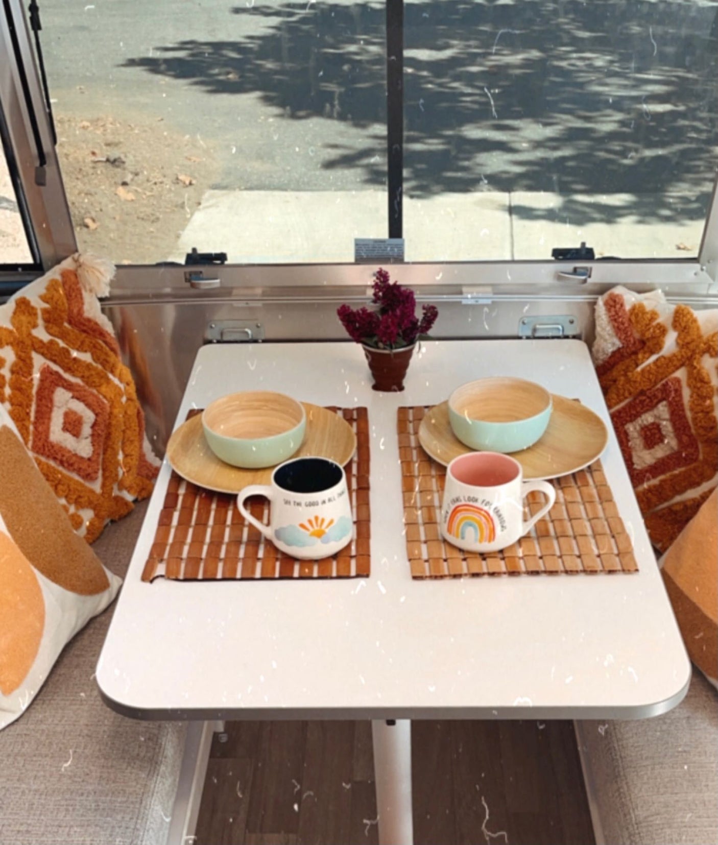 Cozy RV rental dining space with two settings, wood plates, mugs on bamboo mats, and a scenic window view.