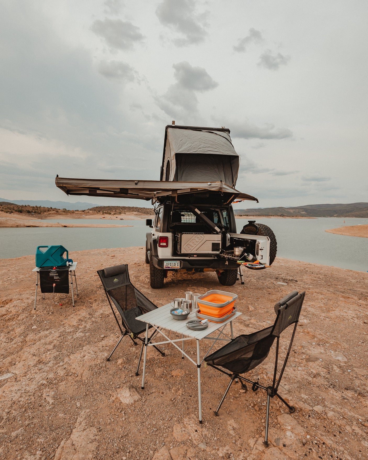 Explore by lake with this overlanding Jeep rv rental, featuring a rooftop tent, awning, and full camp kitchen setup.