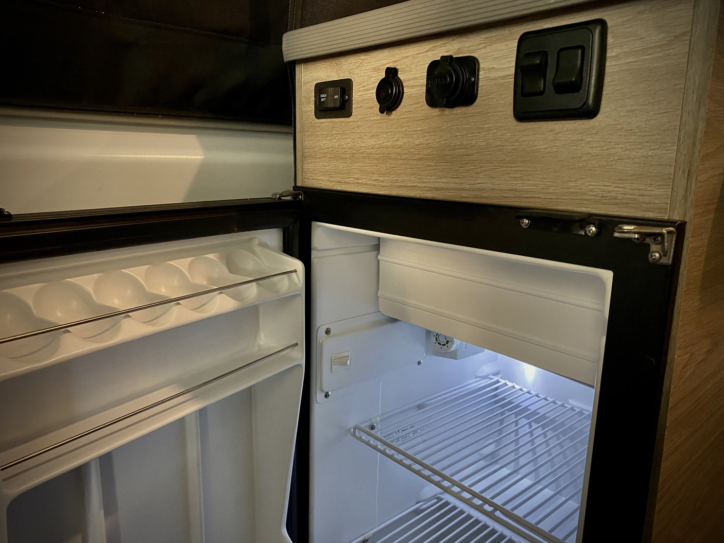 RV rental interior: Open mini-fridge with egg storage and illuminated shelves, plus overhead control panel with power outlets.