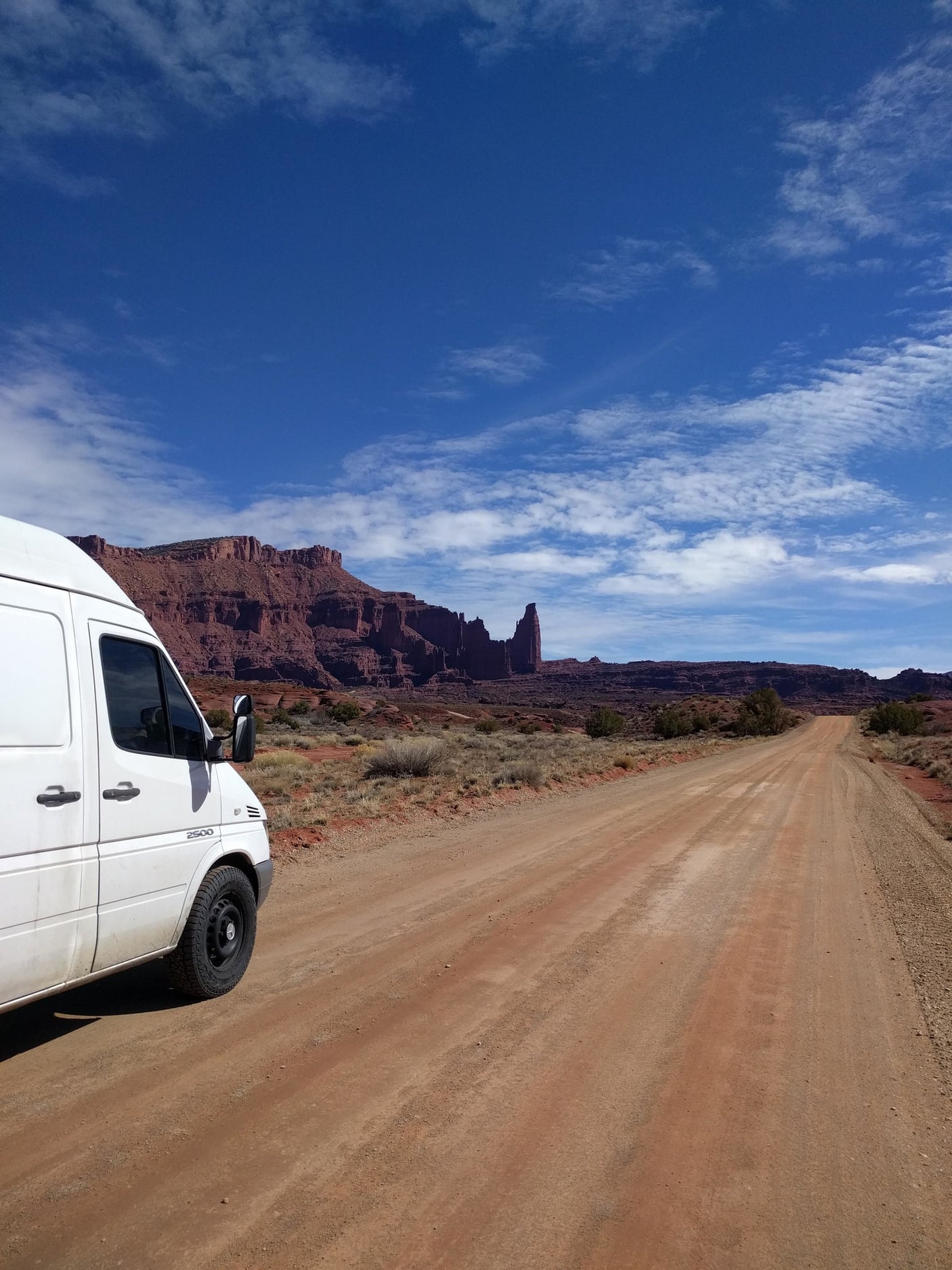 A white camper van, perfect for an RV rental, parked on a red dirt road stretching towards striking red rock mountains under a blue sky.