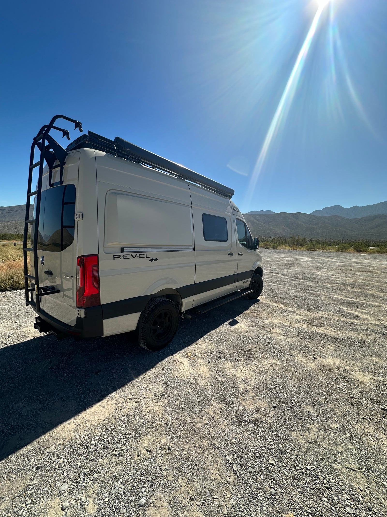 RV rental: Winnebago Revel 4x4 camper van parked on gravel, ready for adventure under a bright, sunny sky with mountains.