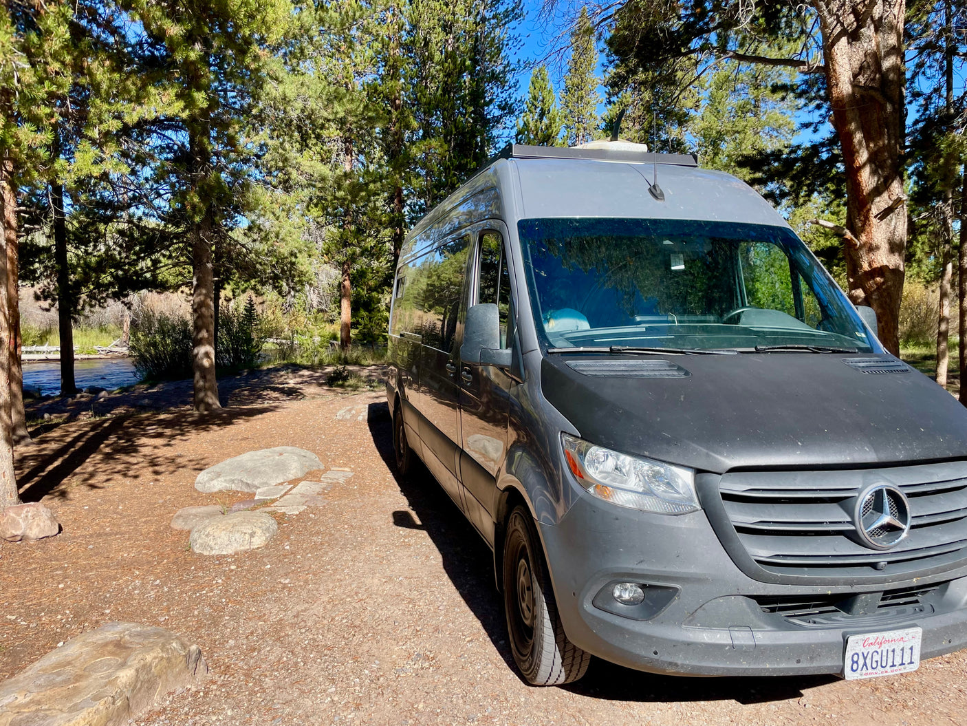Enjoy nature with this grey Mercedes-Benz RV rental parked by a peaceful river among pine trees.
