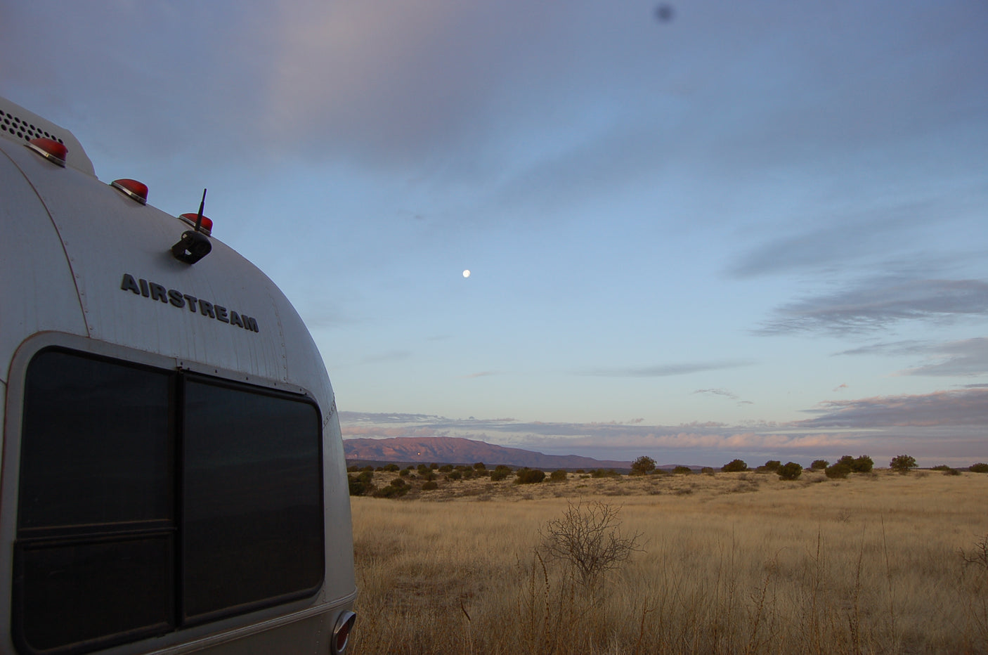 An Airstream RV rental is seen from behind, parked in a vast desert landscape at dusk under a faint moon.