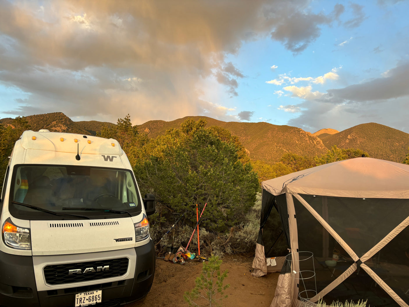 An RV rental camper van and a large screen tent are set up in a mountain campsite at sunset with clouds above.