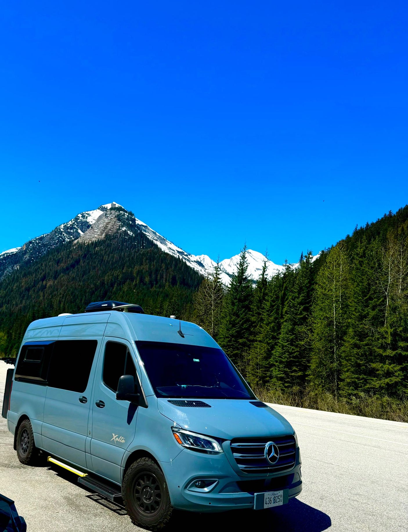 Adventure awaits! This light blue Xalta RV rental is parked with snow-capped mountains and green forests in the background.