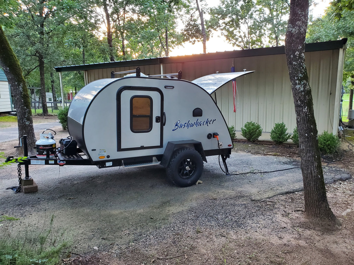 A compact Bushwhacker teardrop camper, ideal for an RV rental, is parked at a wooded campsite with its hatch open.
