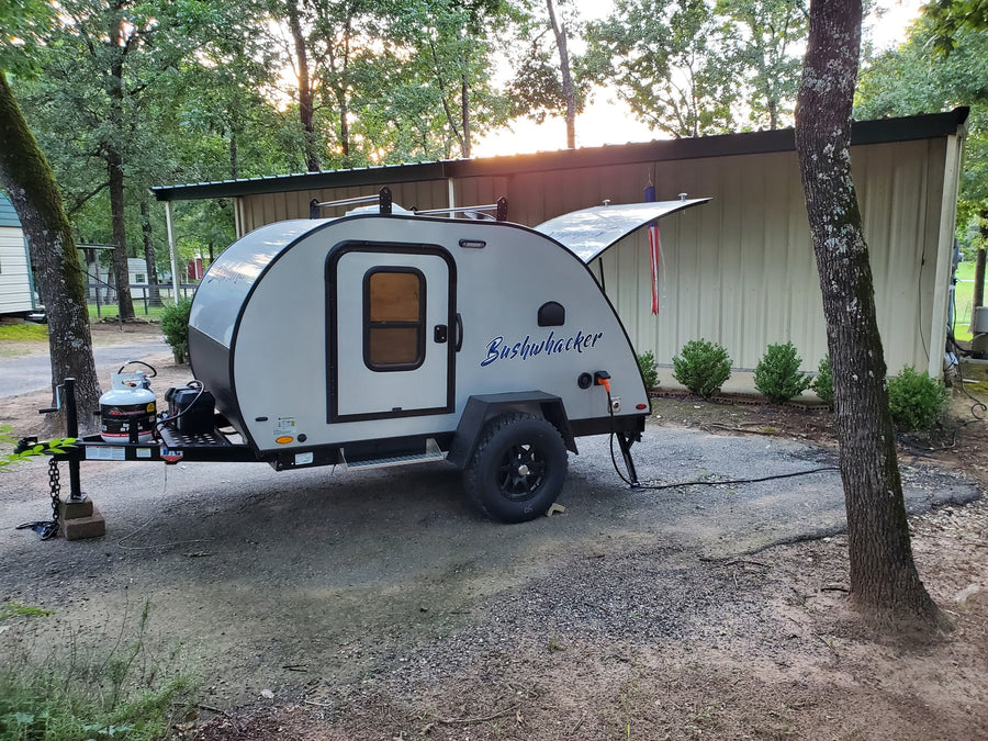 A compact Bushwhacker teardrop camper, ideal for an RV rental, is parked at a wooded campsite with its hatch open.