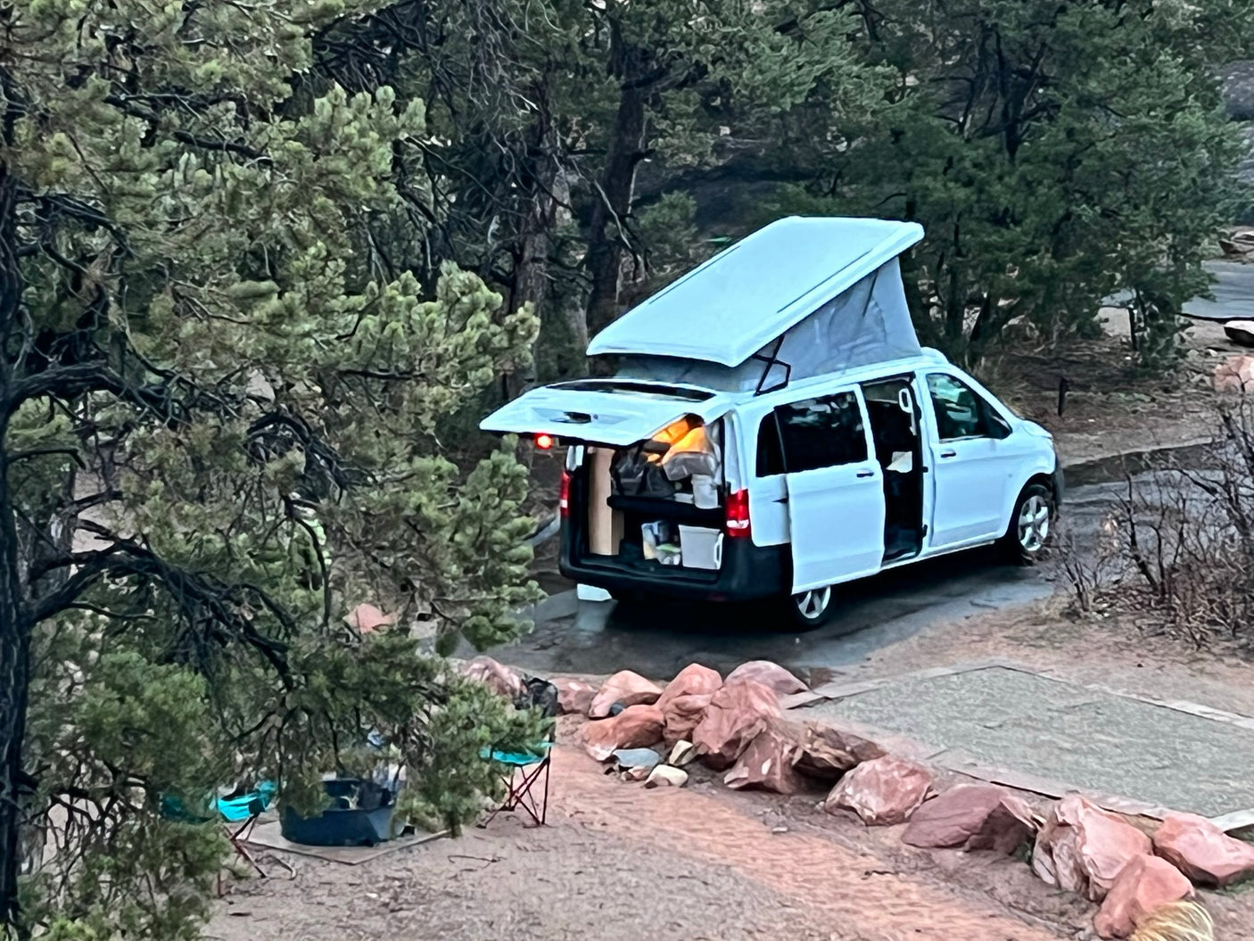 A white pop-top RV rental camper van is parked with doors open at a wooded campsite, ready for an outdoor adventure.