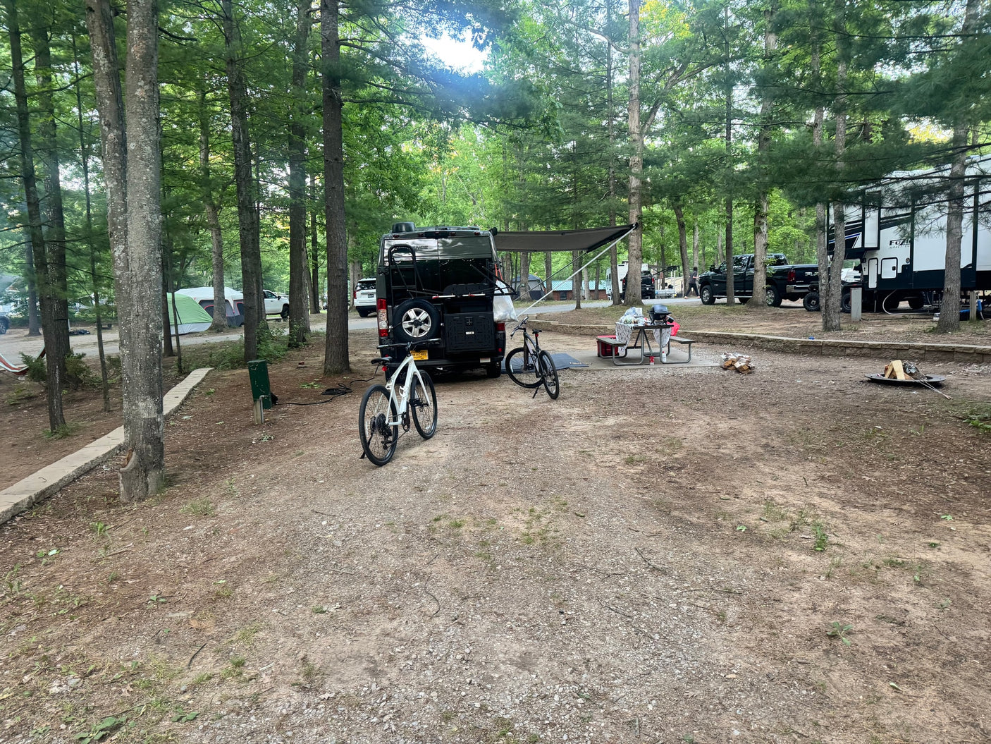 Campsite with a black van-style rv rental, bikes, picnic table, grill, and fire pit under trees.