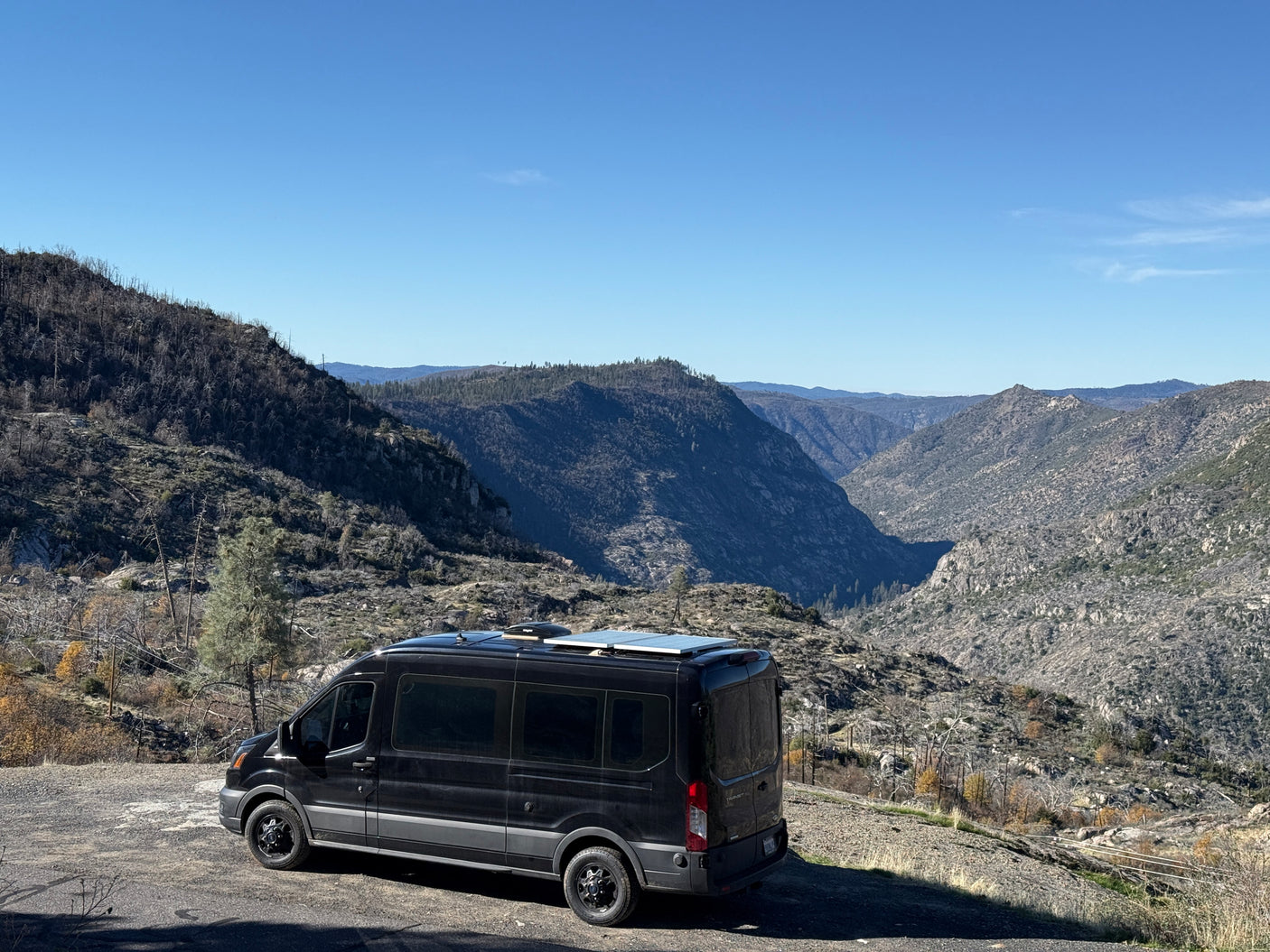 A black camper van (RV rental) parked on a dirt road overlooking a vast, rugged mountain valley with a clear blue sky.