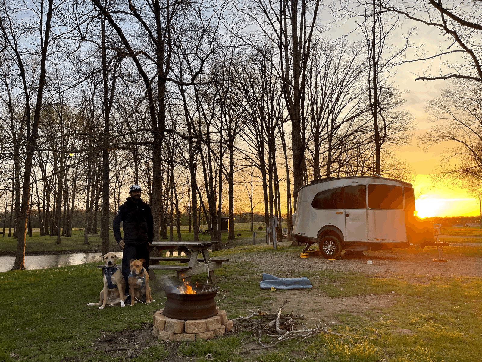 Camping bliss! A person and two dogs by a campfire, with a modern travel trailer RV rental glowing in the sunset.