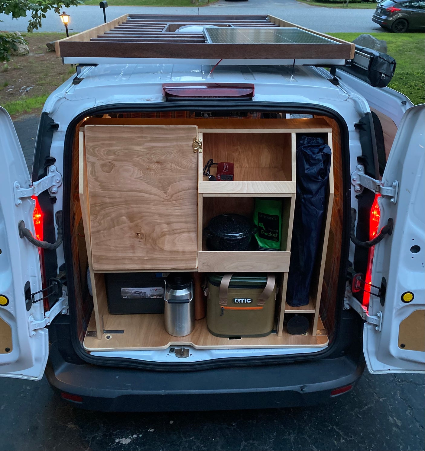 Back of an RV rental van showcasing a DIY camper conversion with organized wood storage, solar panel, and gear.