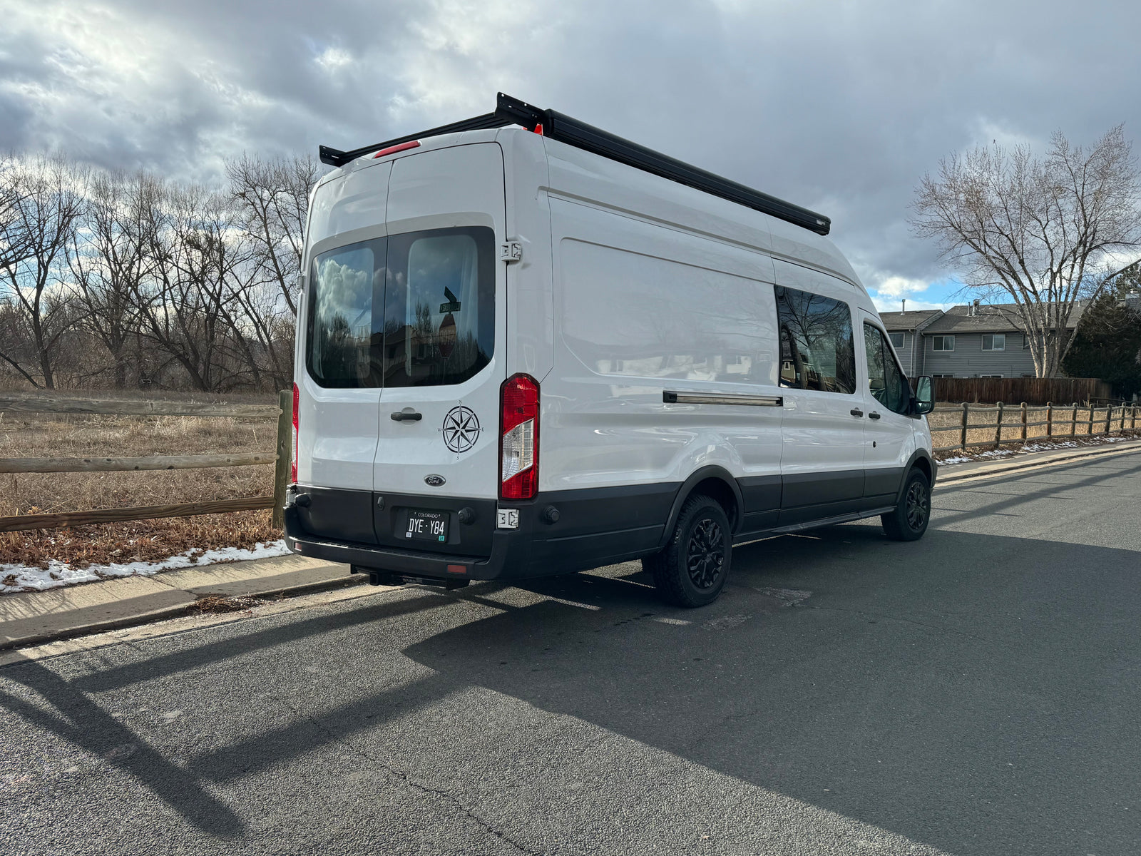 White Ford Transit camper van for RV rental, parked outdoors. Features roof rack, awning & compass decal.