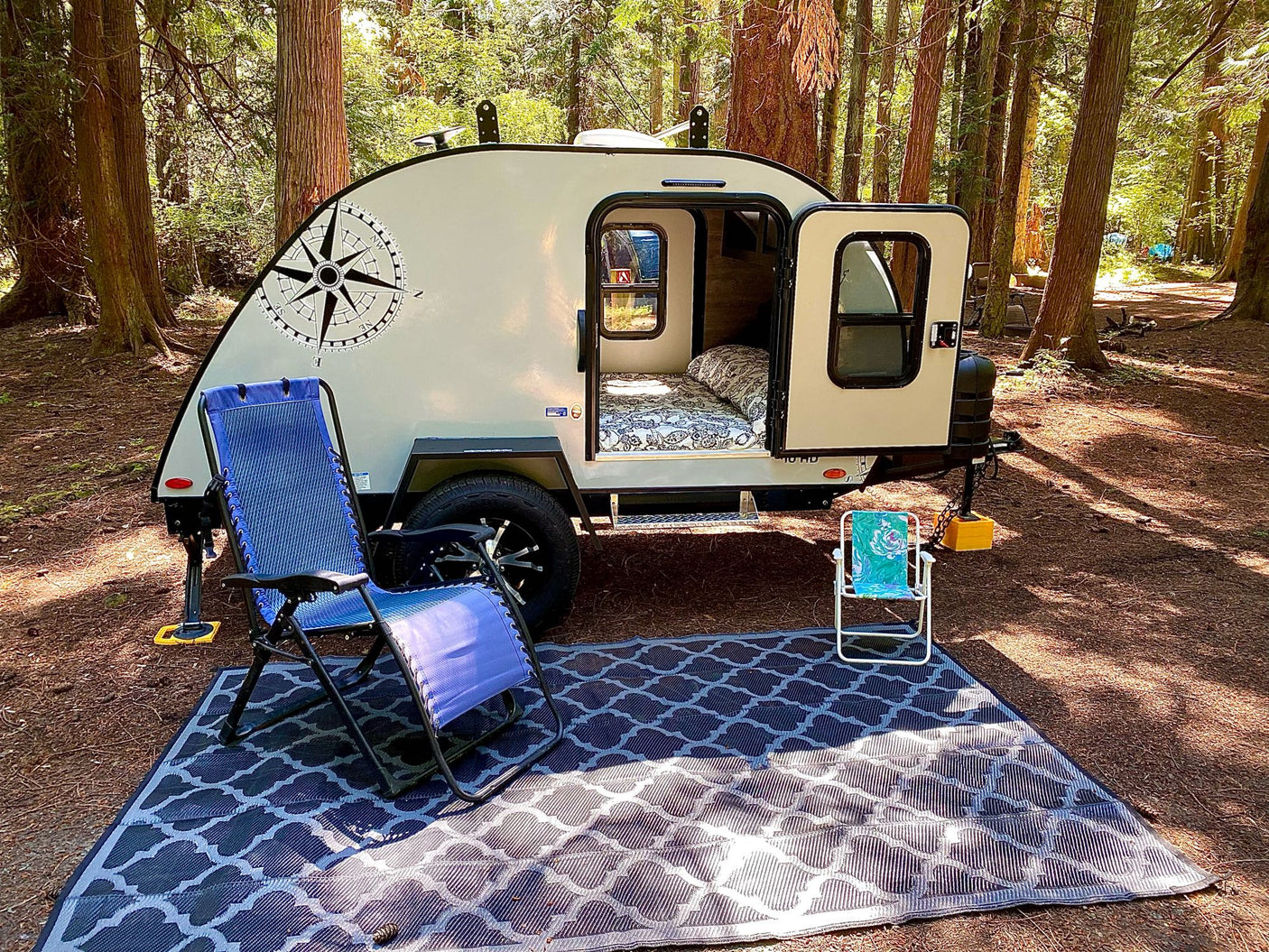 Cozy teardrop RV rental setup in a serene forest campsite. Features open door, bed, chairs, and outdoor mat.