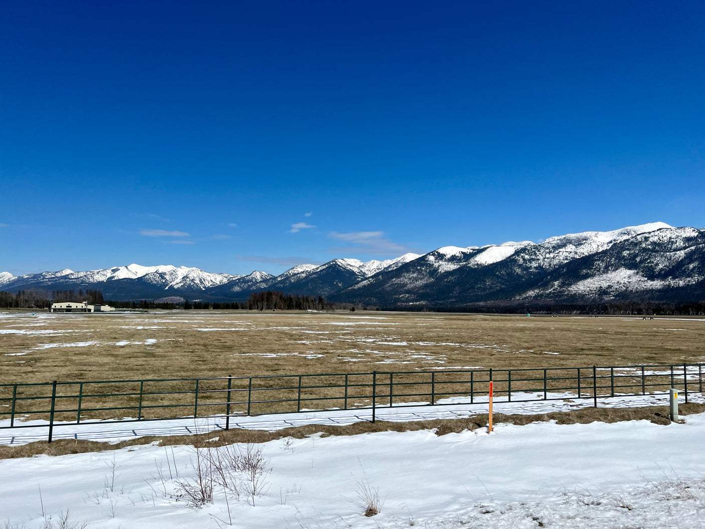 Snowy mountains frame a vast field, melting snow, and a fence under a blue sky. Perfect for an RV rental trip.