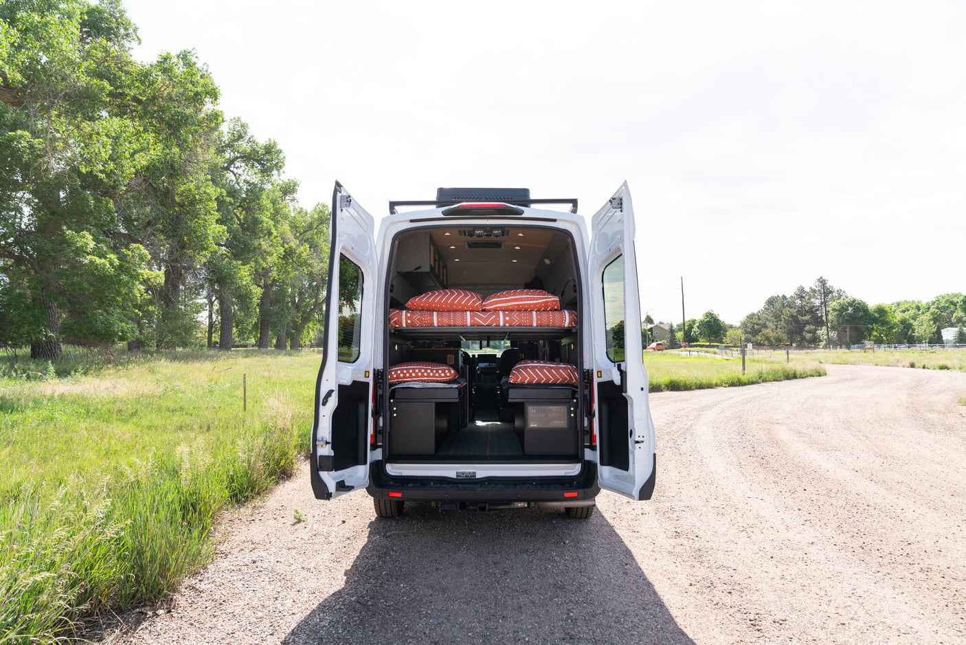 This custom RV rental camper van features dual bunk beds, seen with its back doors open on a gravel road, inviting adventure.