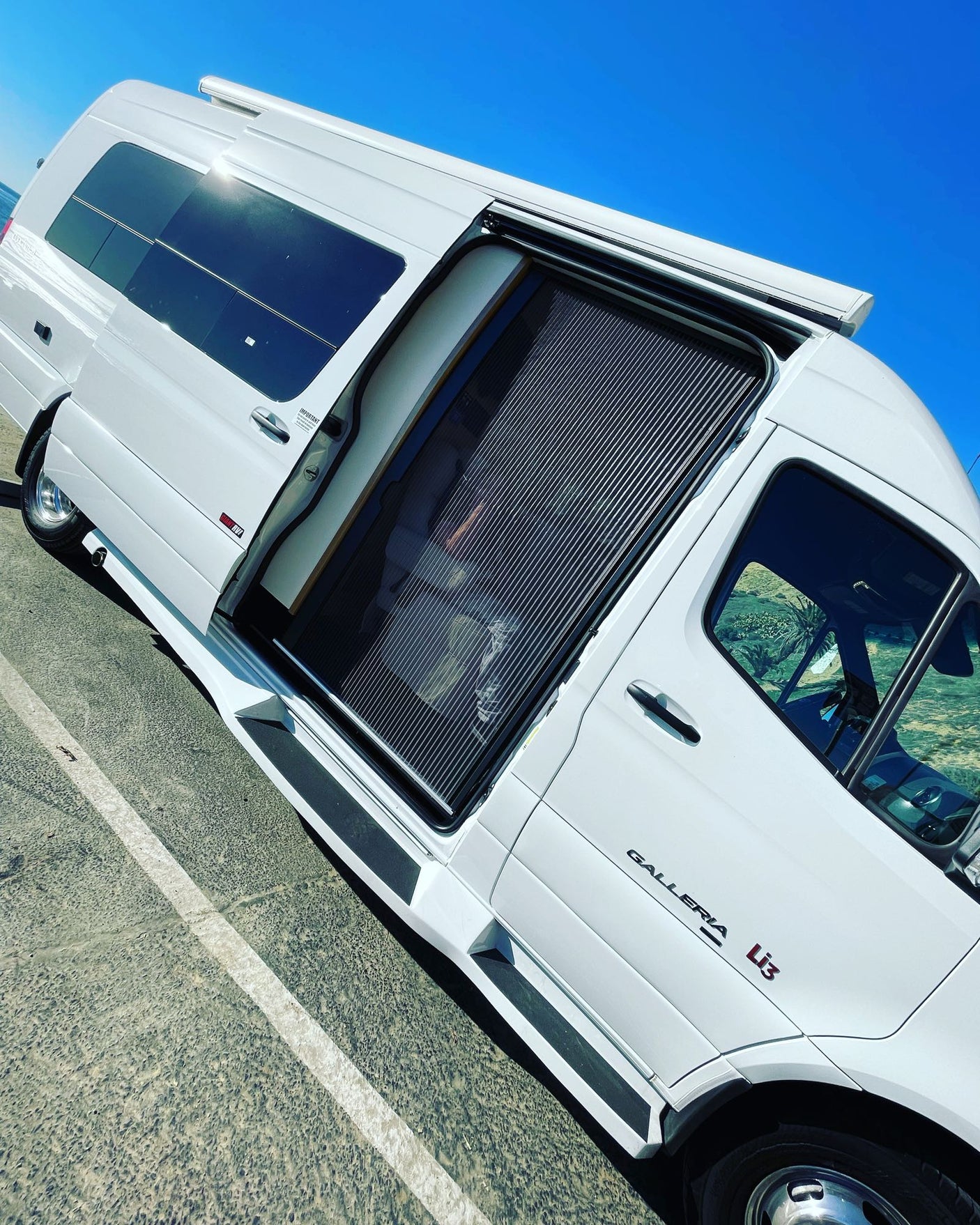 This white Galleria Li3 Class B RV rental, a van-style motorhome, with its side door open revealing a screen, under a clear blue sky.