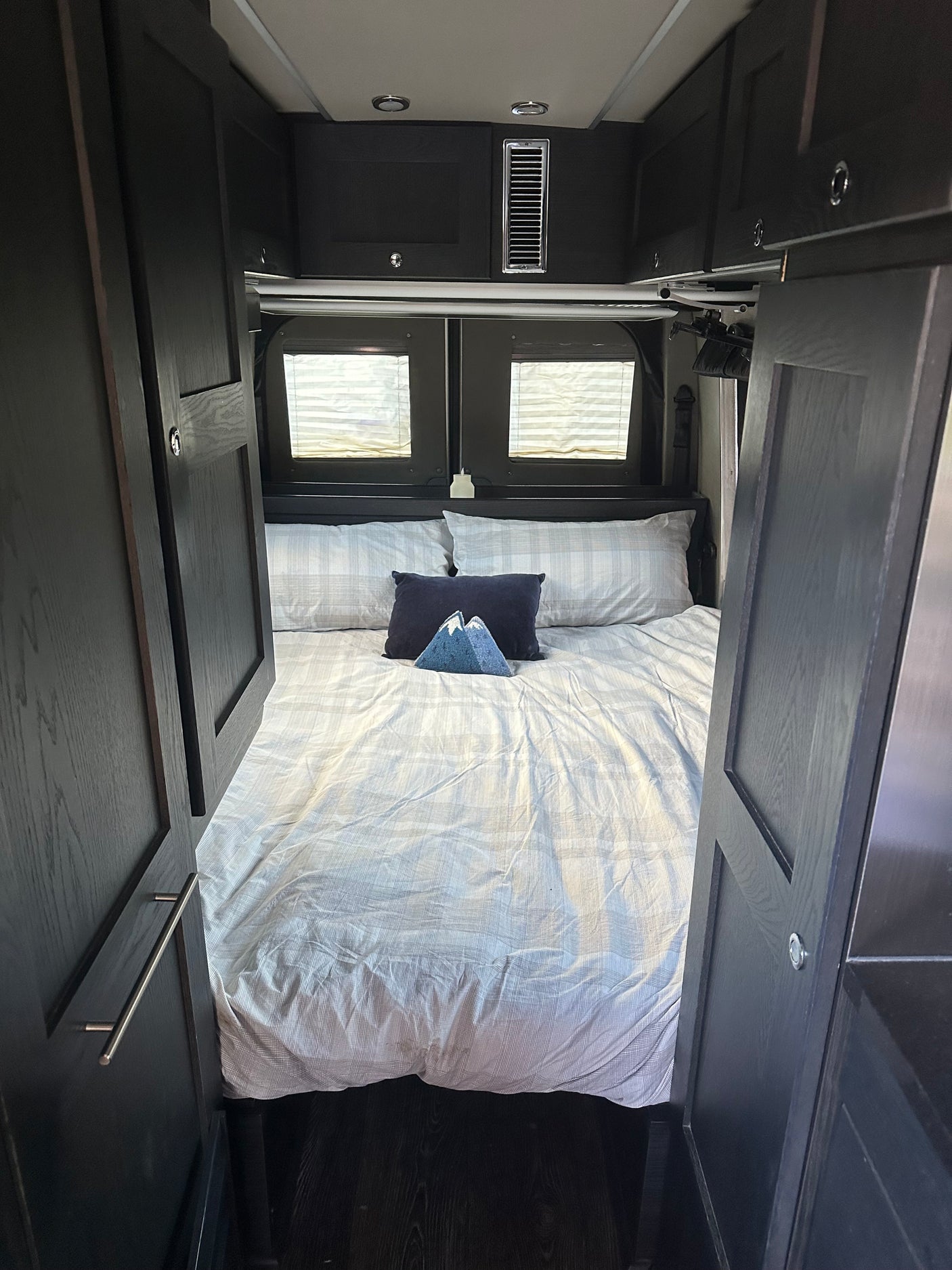 Step into this comfortable RV rental bedroom. Features a large bed with striped bedding, dark cabinets, and rear windows.