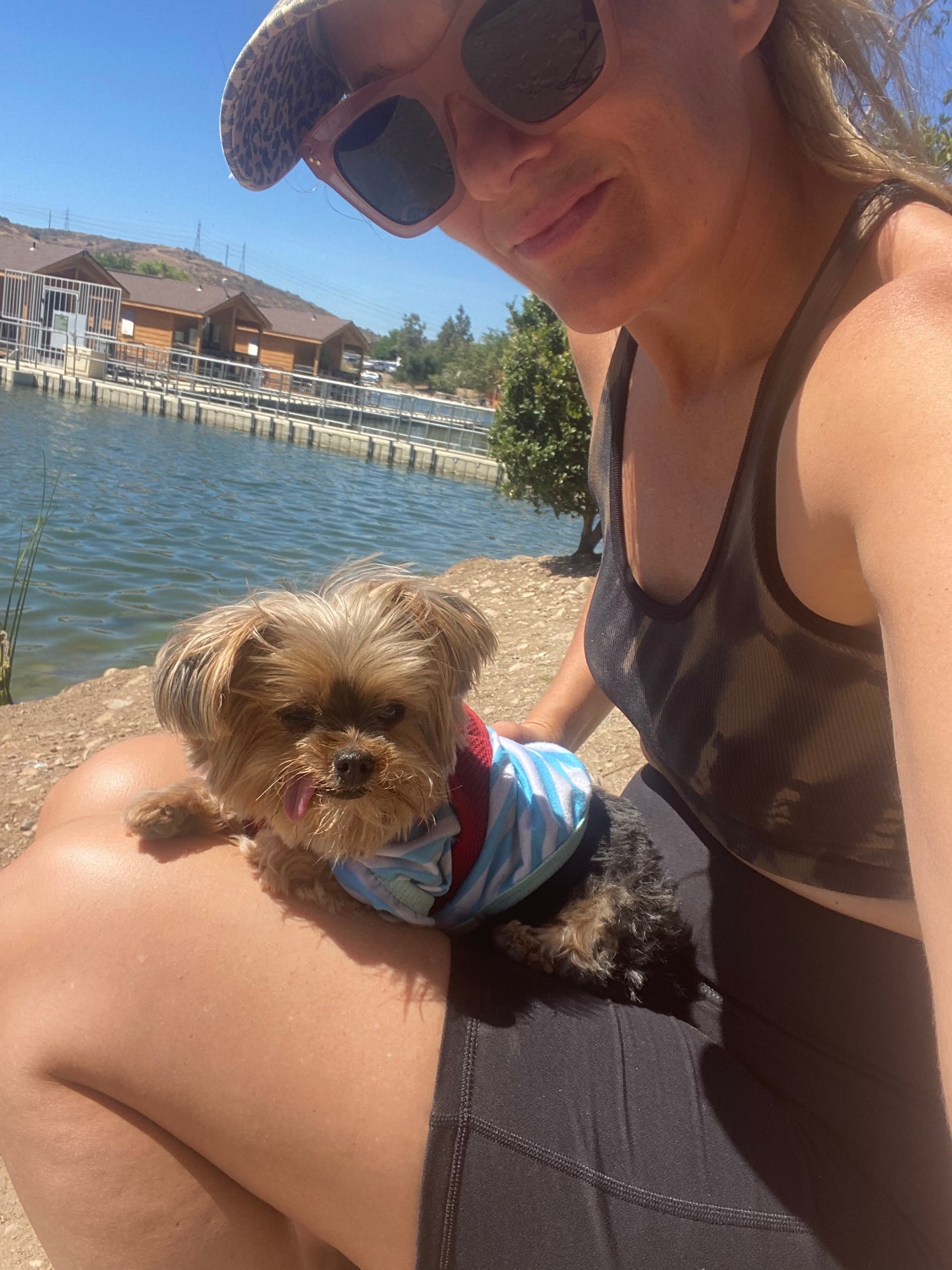 A person in sunglasses holds a small dog by a scenic lake, a relaxing scene ideal for an RV rental trip.