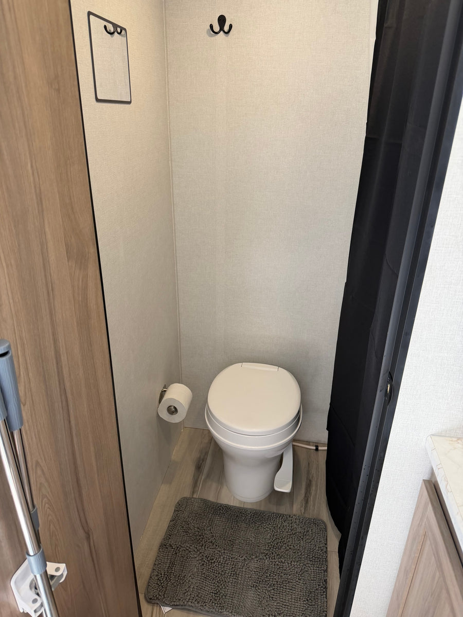 View of a compact RV rental bathroom, showing a white toilet, gray mat, and black shower curtain against light-textured walls. Includes a toilet roll and hooks.