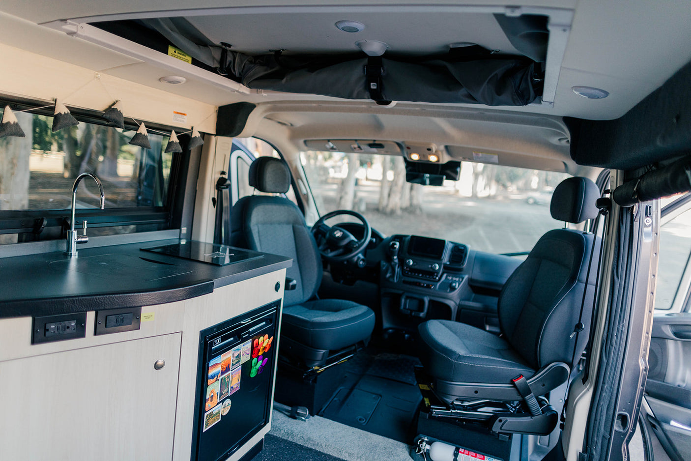 Inside an RV rental: a modern kitchenette with a sink and stovetop, a mini-fridge, front cabin seats, and overhead storage.