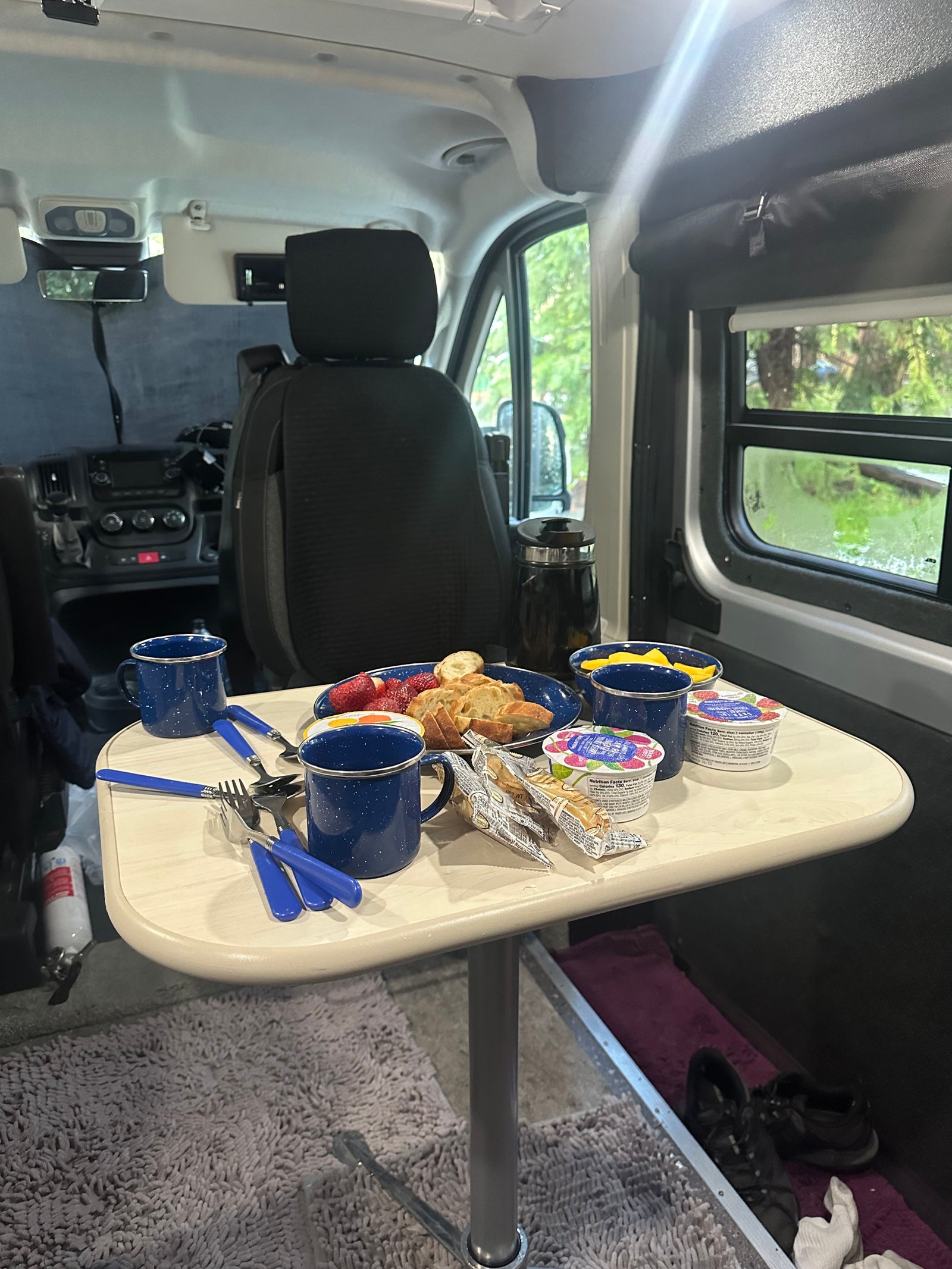 Cozy RV rental breakfast inside a camper van, featuring fruit, bread, and coffee with nature views.