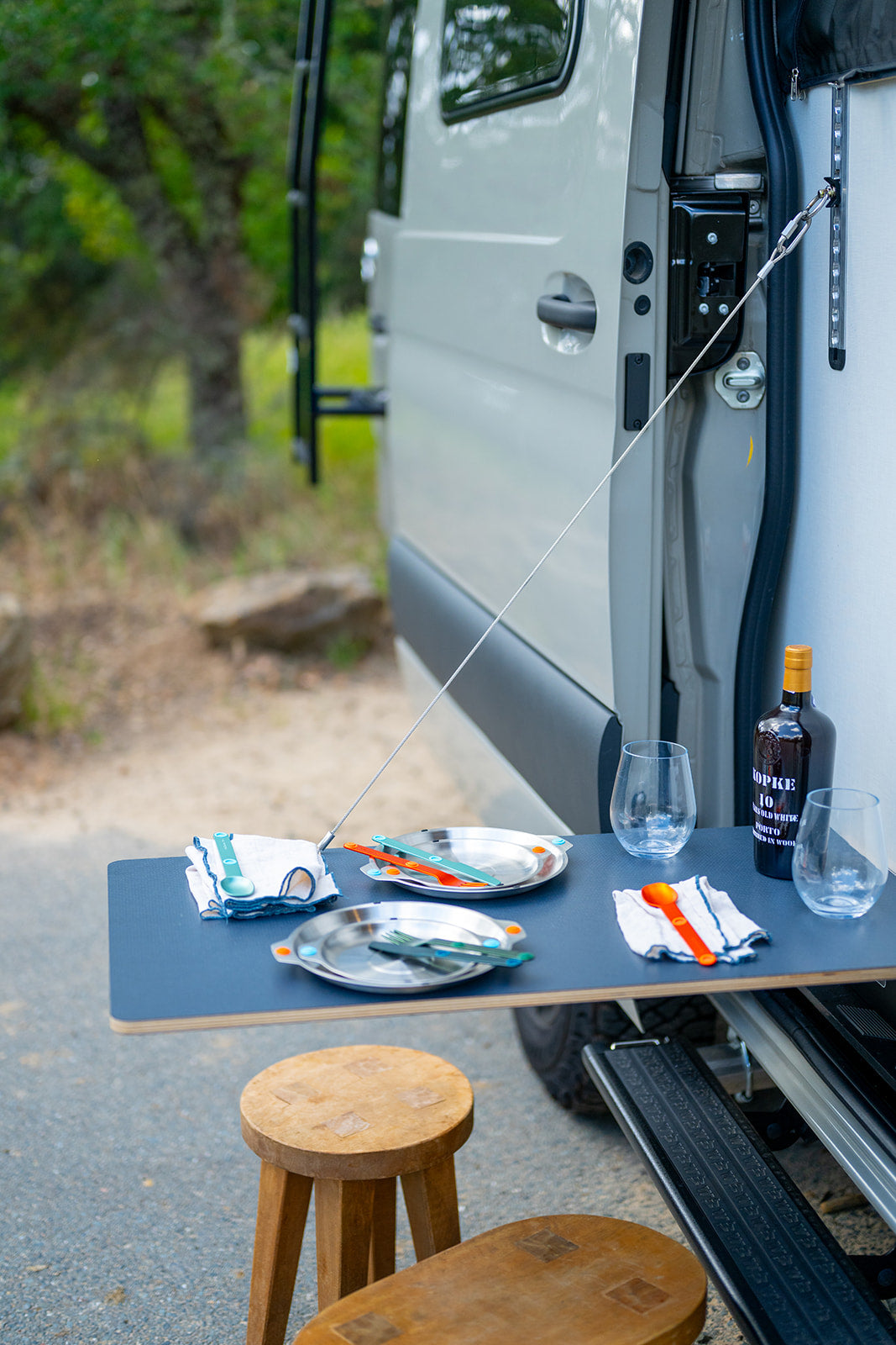 RV rental outdoor dining: Camper van's side table with plates, colorful cutlery, glasses, and wine bottle for a camp meal.