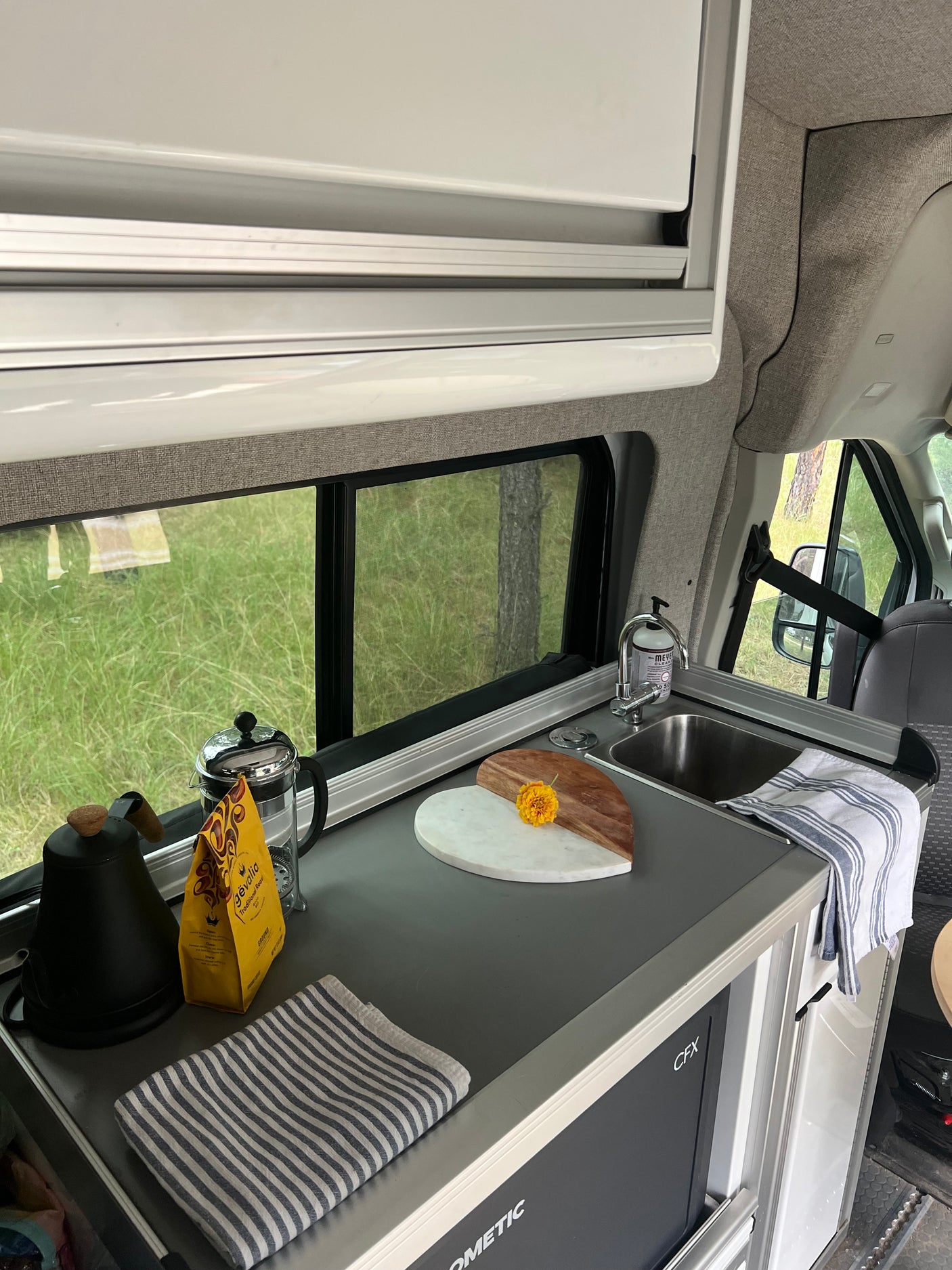 Inside an RV rental: a well-equipped kitchenette with coffee, sink, Dometic fridge, and a green outdoor view.