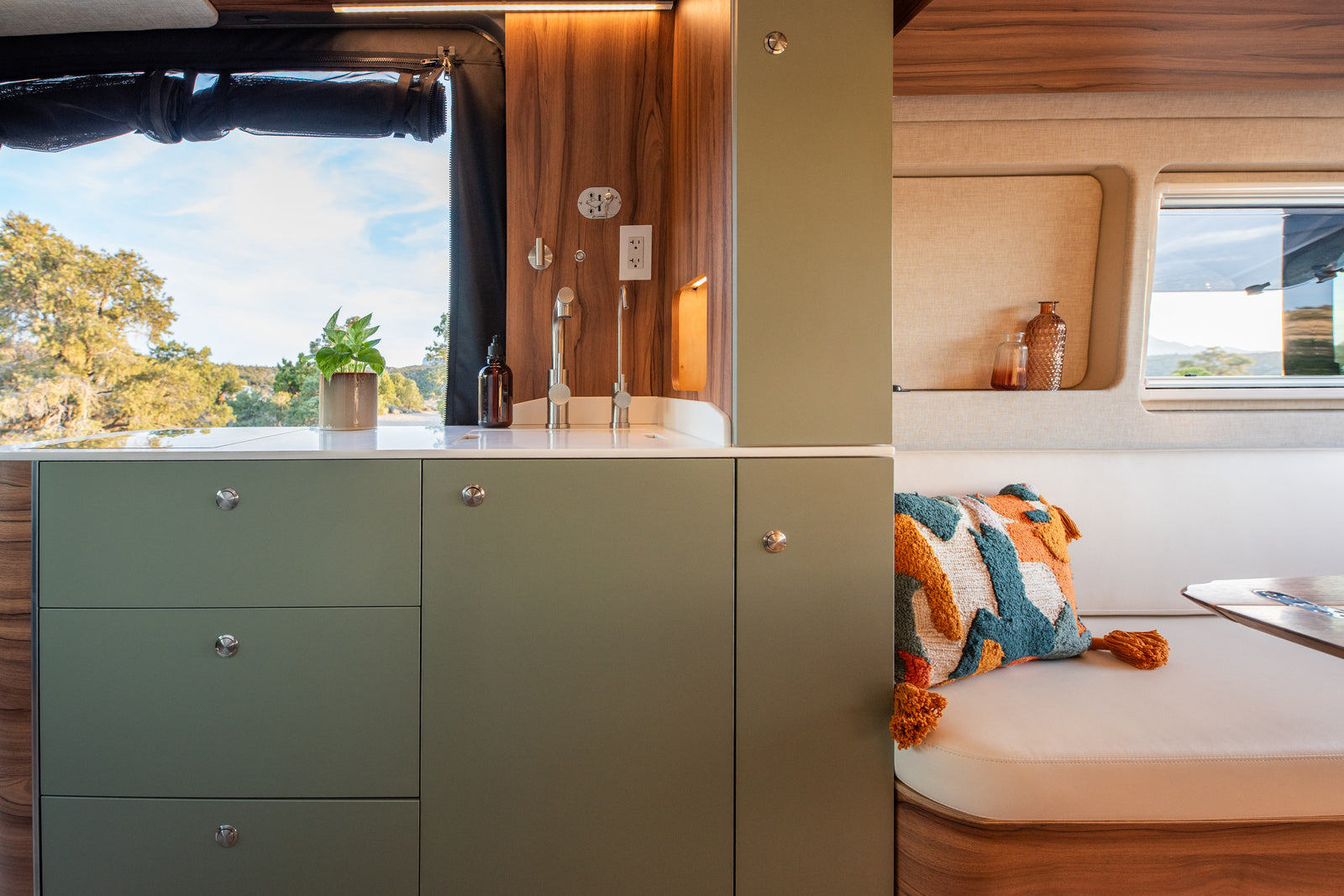 Modern RV rental interior features a sage green kitchen with a sink and a cozy dining nook with a vibrant cushion, framed by scenic outdoor views.