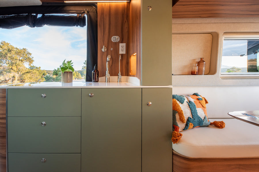 Modern RV rental interior features a sage green kitchen with a sink and a cozy dining nook with a vibrant cushion, framed by scenic outdoor views.