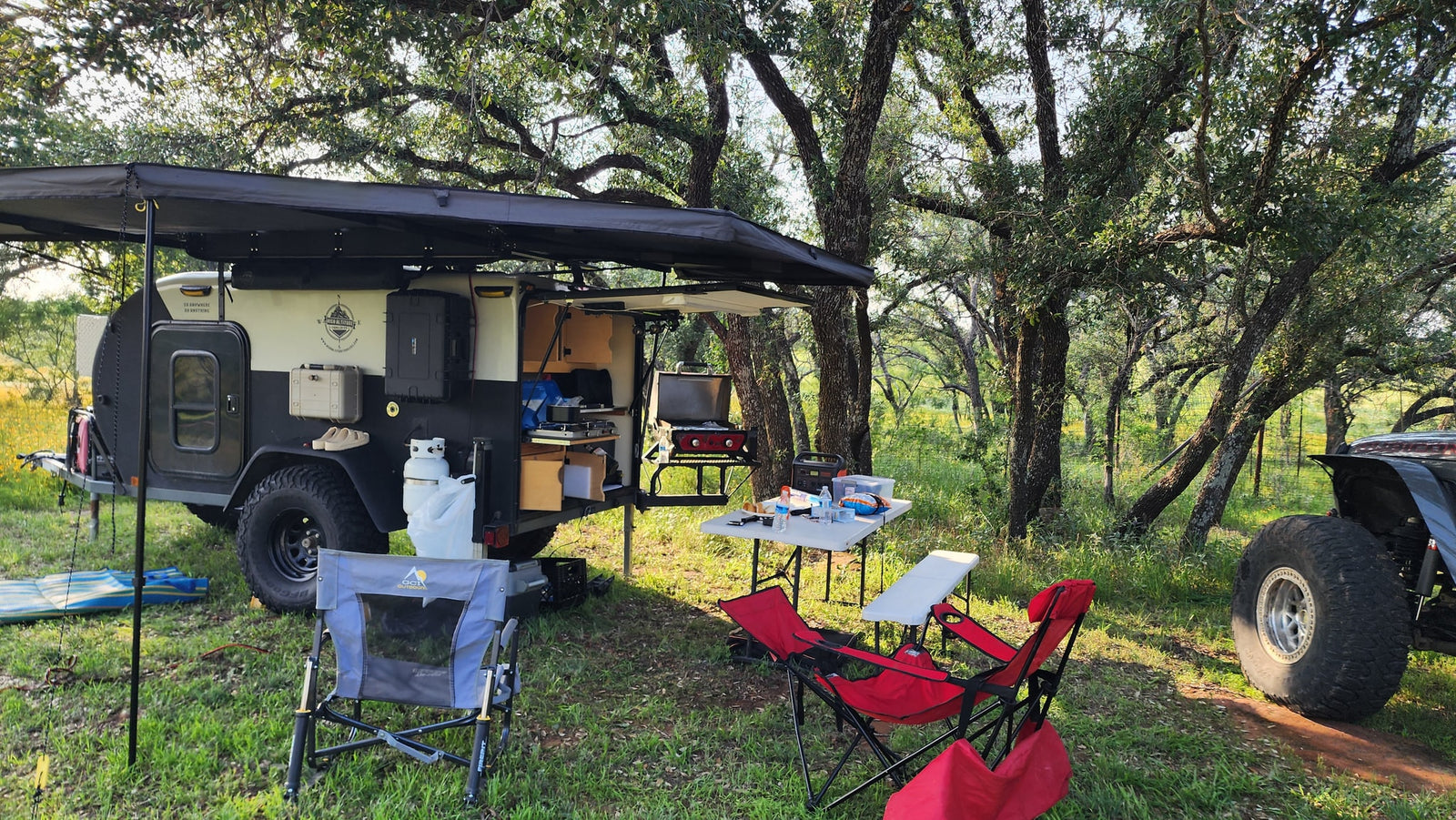 This overland trailer campsite, perfect for an RV rental adventure, features a compact kitchen, grill, and comfy chairs under an awning.