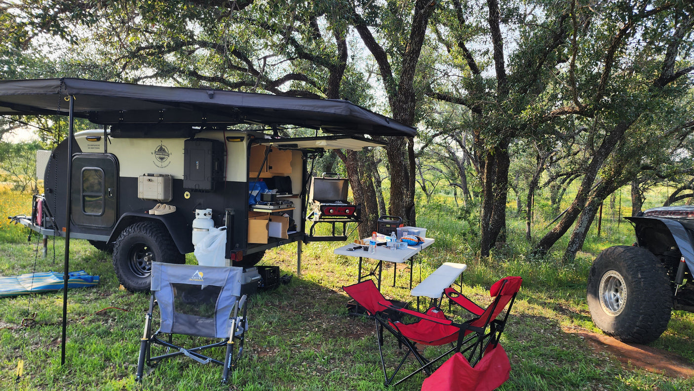 This overland trailer campsite, perfect for an RV rental adventure, features a compact kitchen, grill, and comfy chairs under an awning.