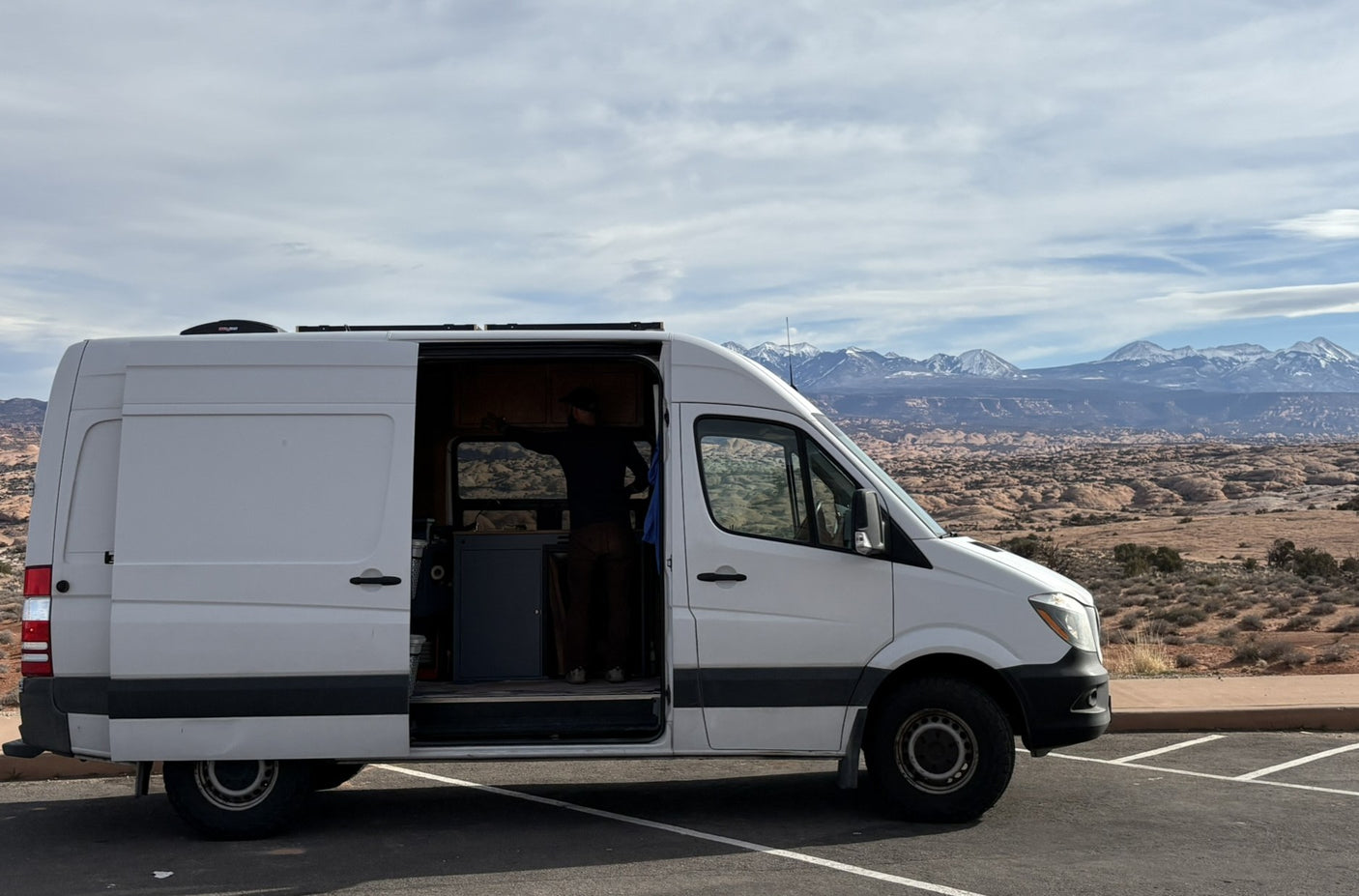 Scenic rv rental adventure: White camper van with open door, person inside, parked amidst desert landscape & snowy mountains.