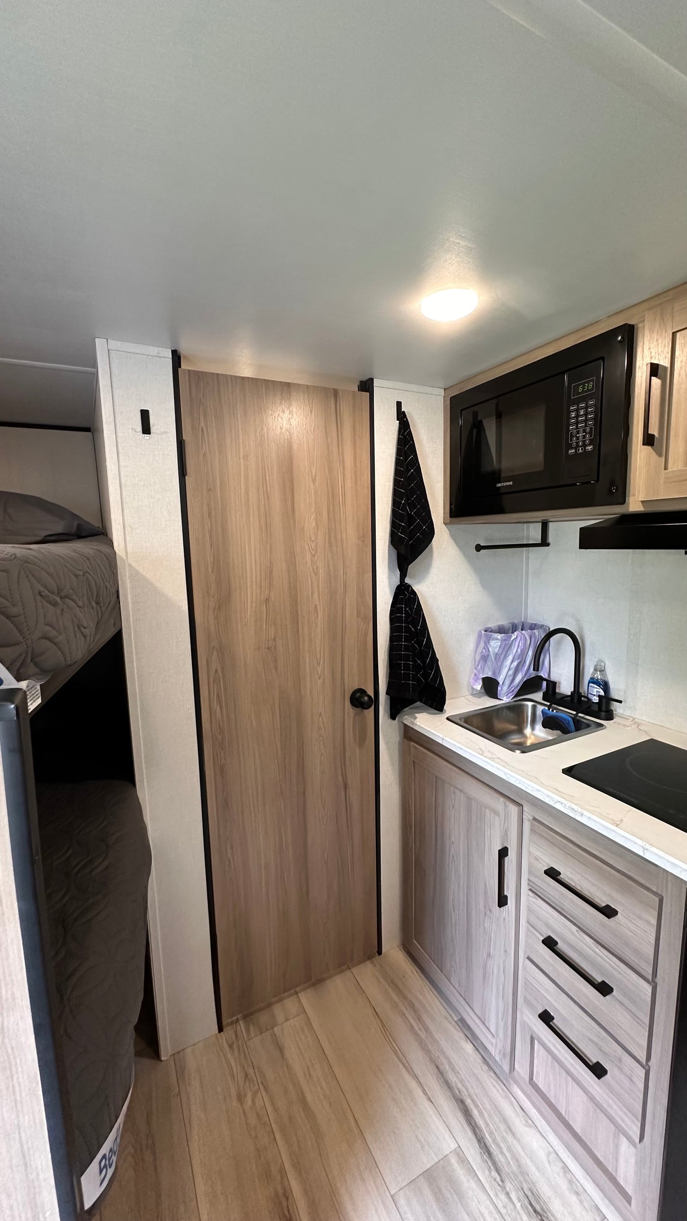 Modern RV rental interior: compact kitchen with microwave, sink, and cooktop, alongside cozy bunk beds. Light wood cabinetry.