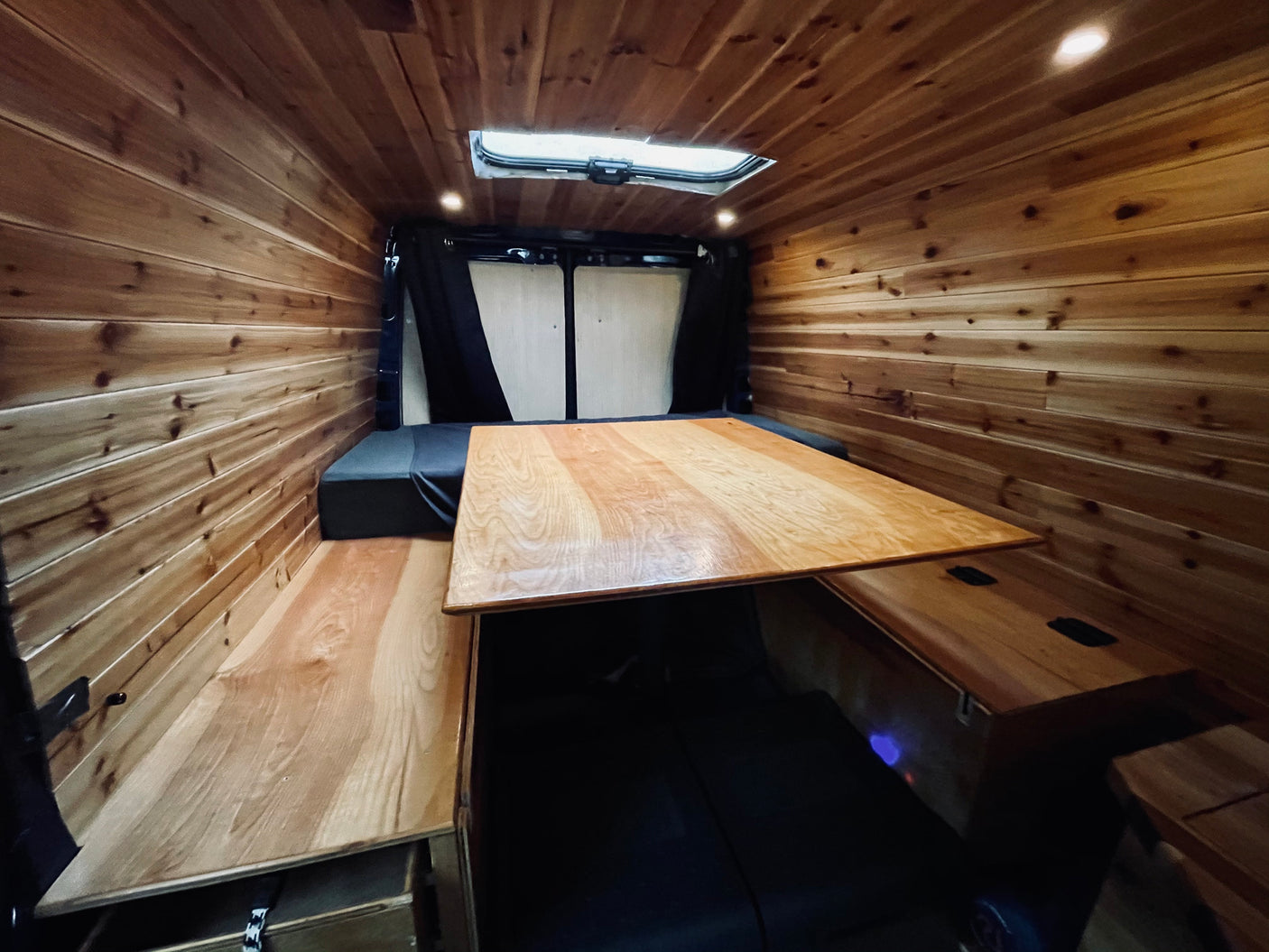 Inside a custom wood-lined RV rental van, featuring a central table and comfortable bench seating.