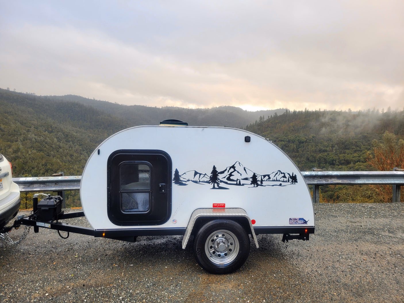 Scenic rv rental: White teardrop trailer with mountain decal, parked on a gravel road overlooking foggy, forested mountains.