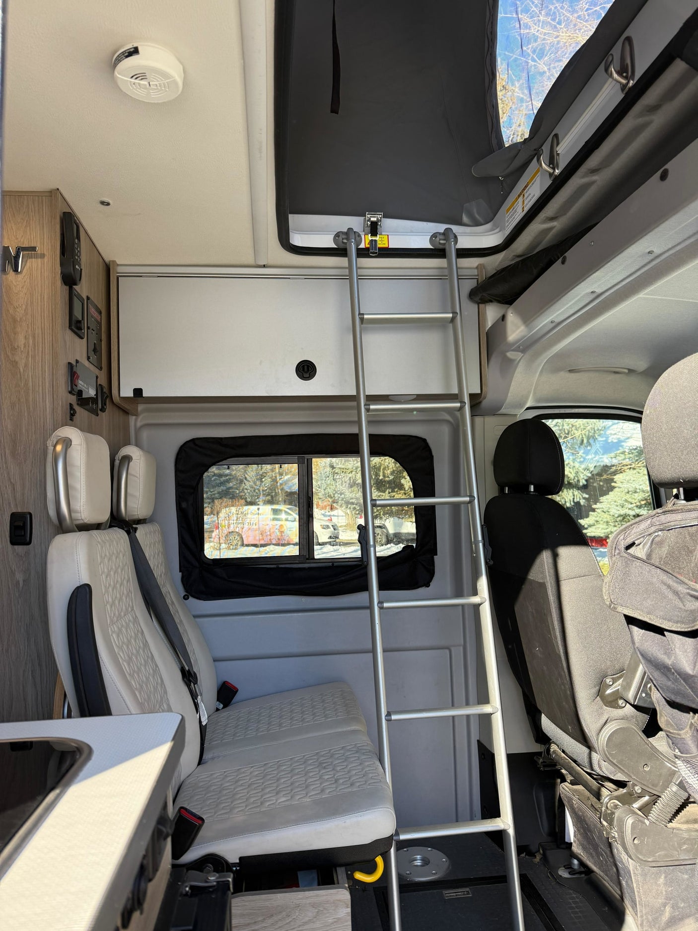 Interior of an RV rental: ladder to a pop-top bunk, passenger seats, wall controls, and a winter window view.