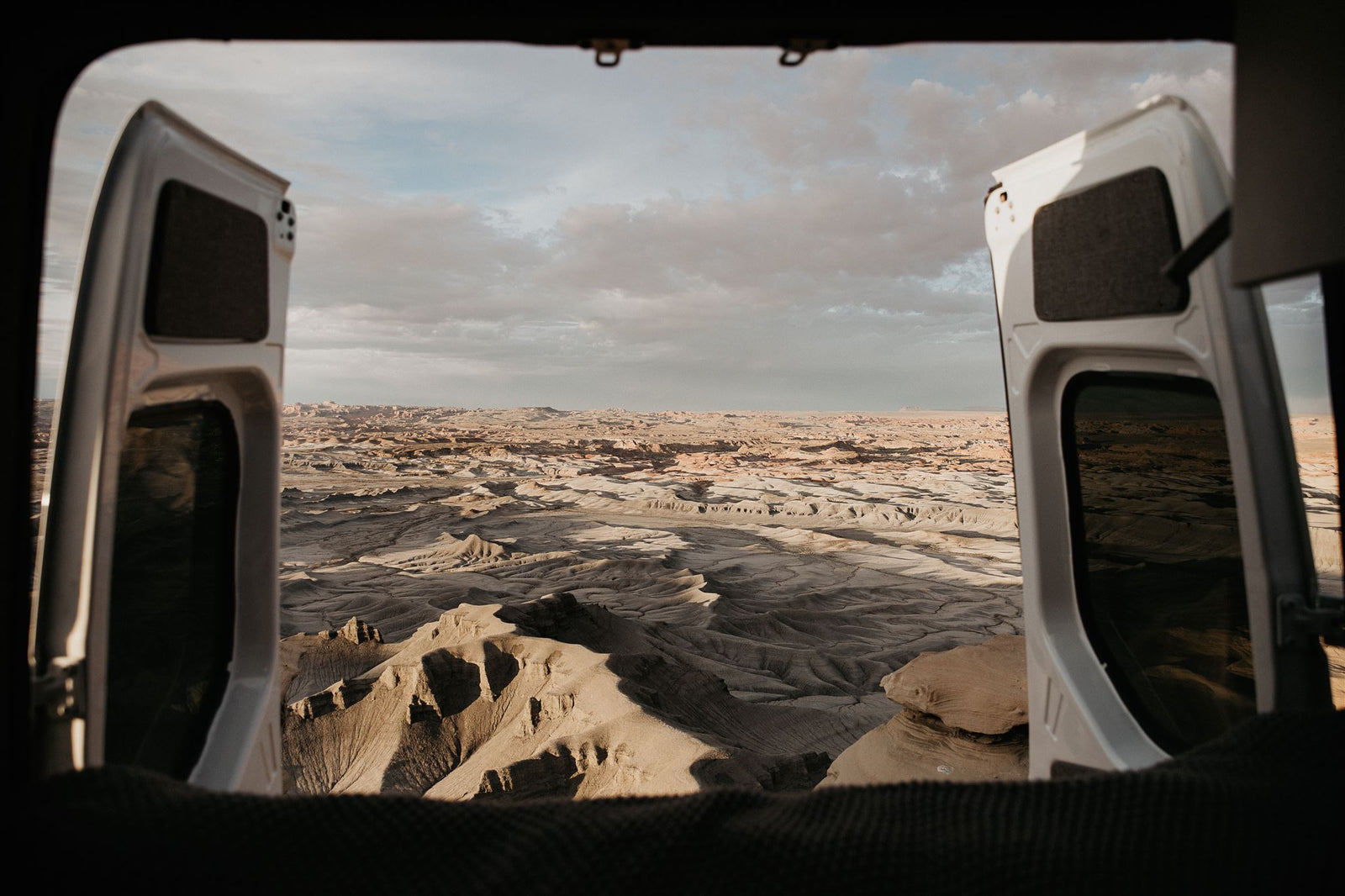 Open RV rental doors frame a stunning desert vista. Textured badlands and distant mesas stretch under a vast, cloudy sky.