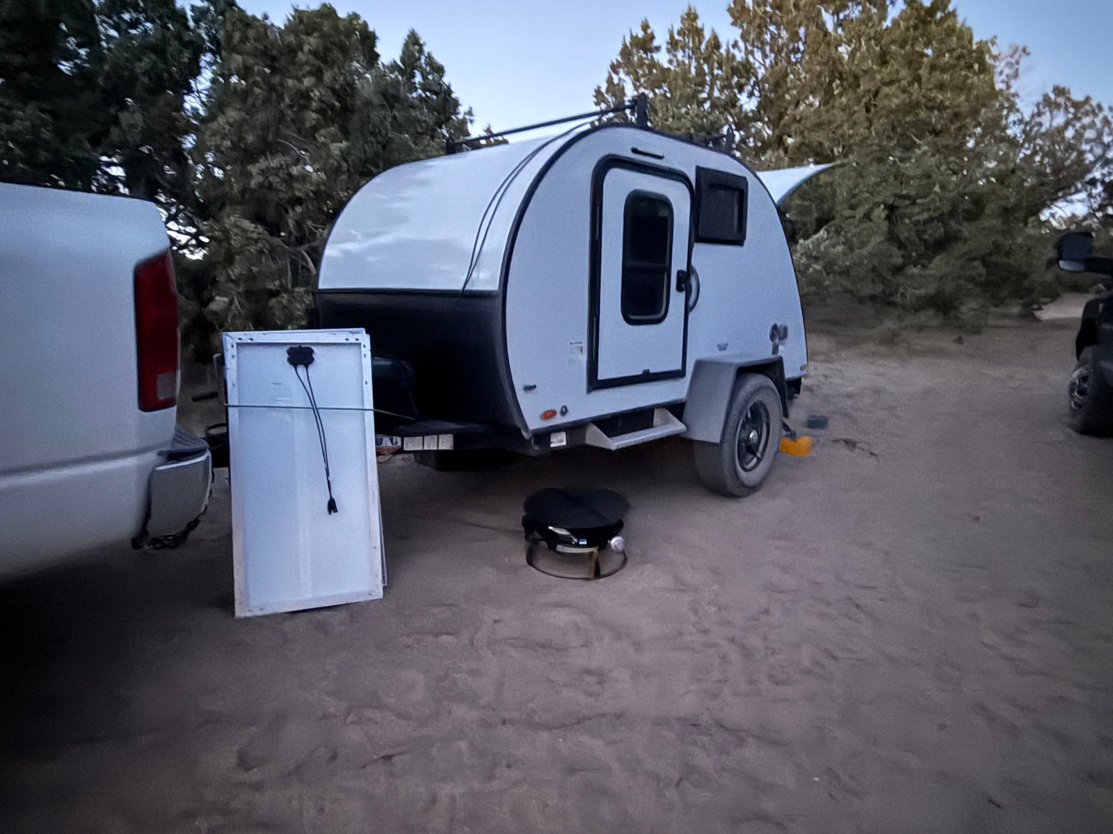 Teardrop RV rental for off-grid camping. Truck, solar panel, and grill set up on dirt amid trees.