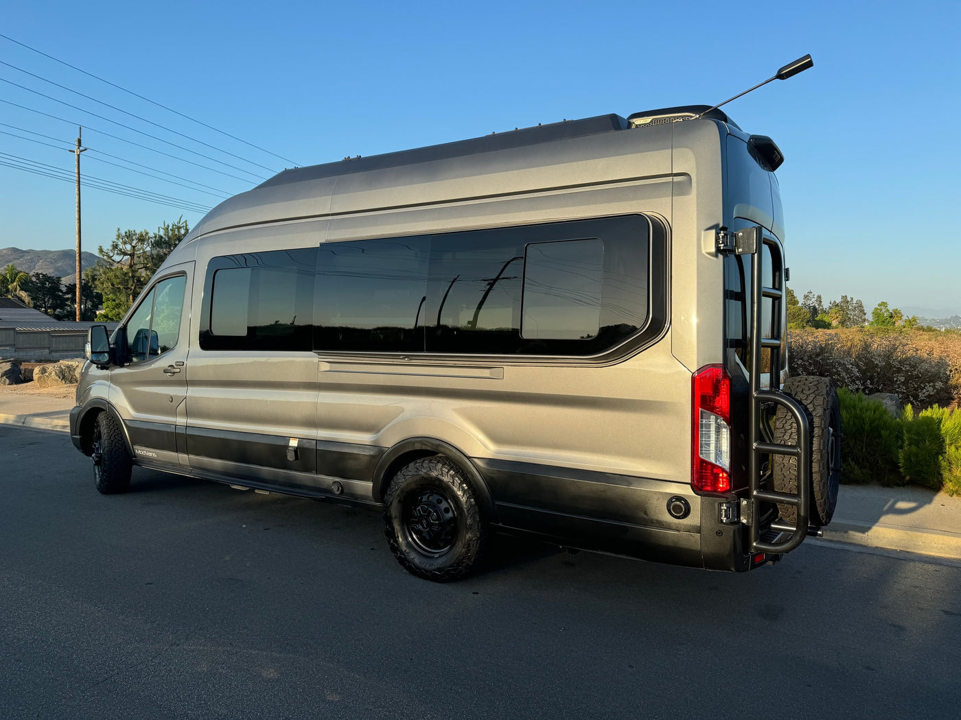 parked on asphalt. Ideal rv rental for your next outdoor adventure.|Close-up of a gray Ford van's front grille with Ford logo & ModVans RV rental license plate (RWT-3467).|Rear view of a gray ModVans Ford Transit rv rental. Features a spare tire — photo 5
