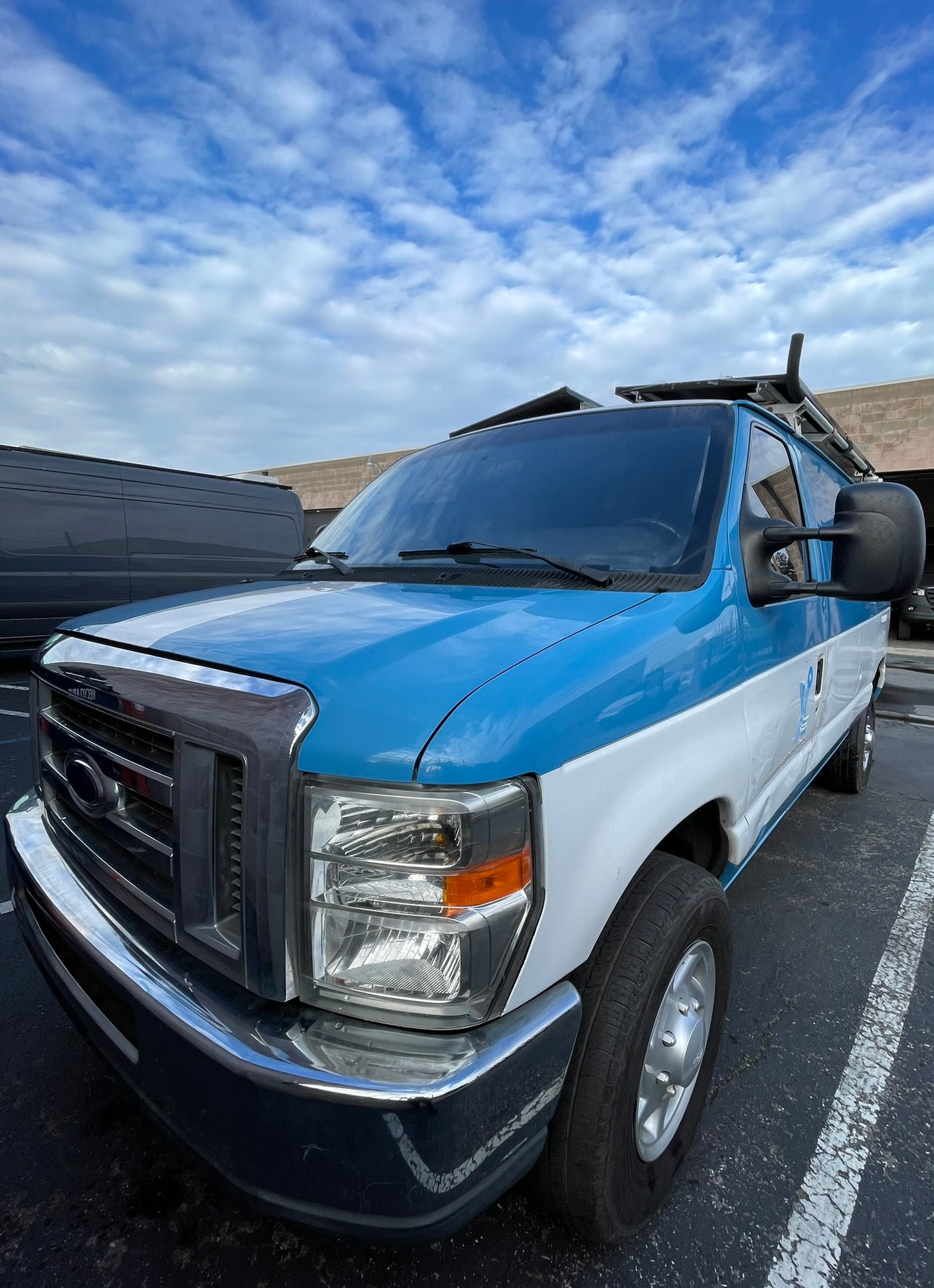 Blue & white van, reminiscent of an RV rental, with a roof rack, parked under a cloudy sky.
