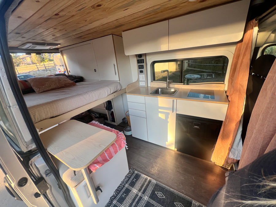 This inviting RV rental offers a raised bed, modern kitchenette with sink & fridge, and controls for an amazing van life.
