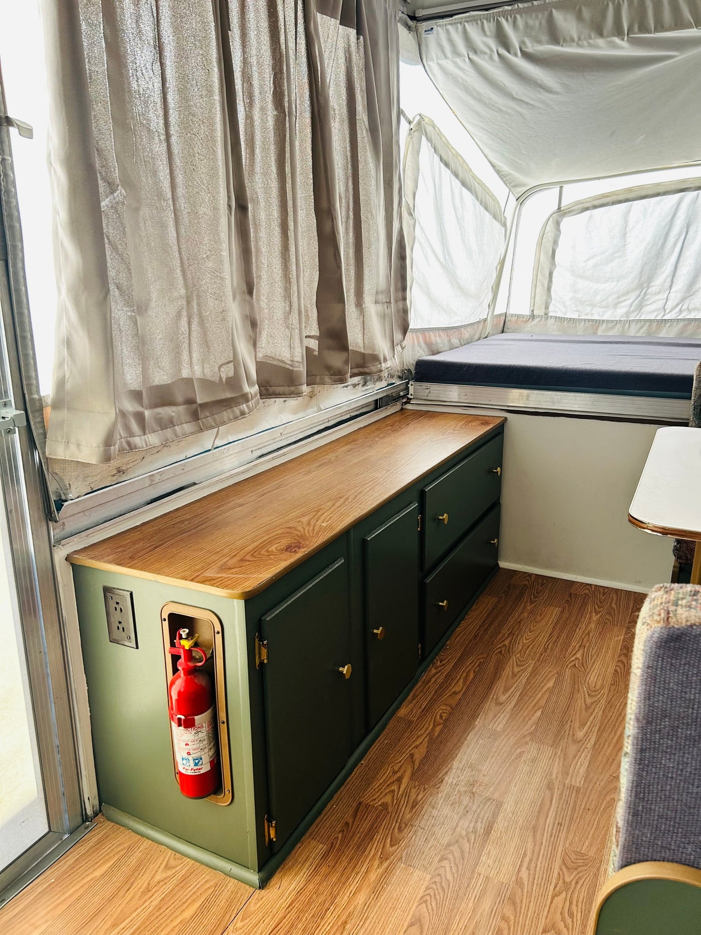 This rv rental showcases custom dark green storage cabinets with a wood top, a bed, and a fire extinguisher, offering functional comfort.