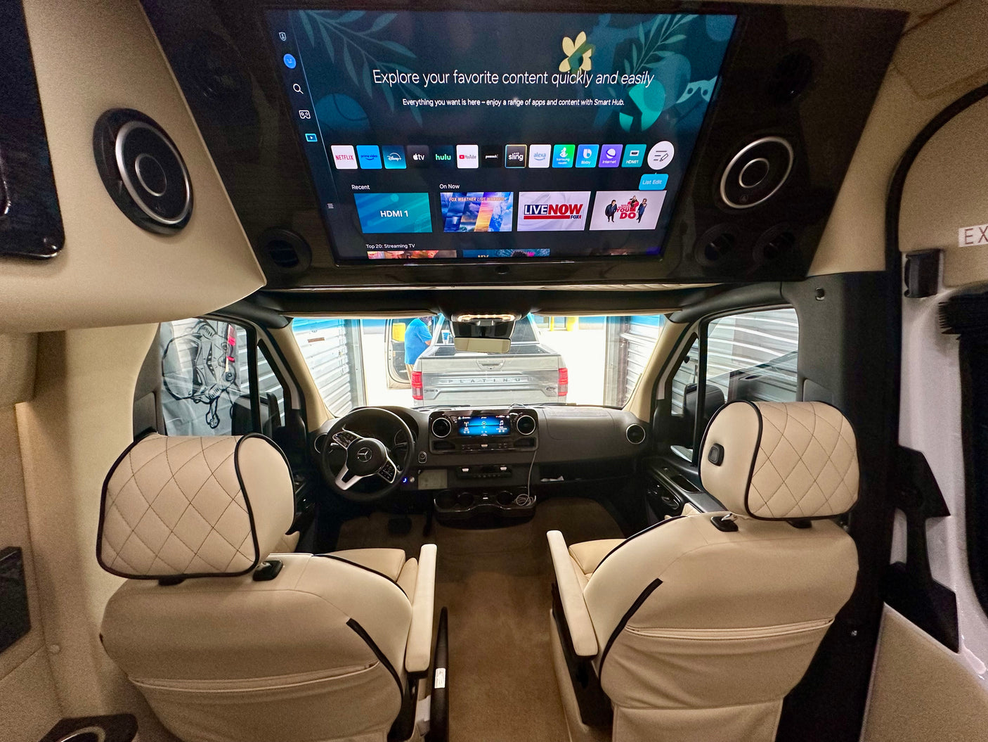 Luxurious RV rental interior, featuring a large overhead Smart TV for entertainment, quilted cream leather seats, and a Mercedes-Benz cockpit.