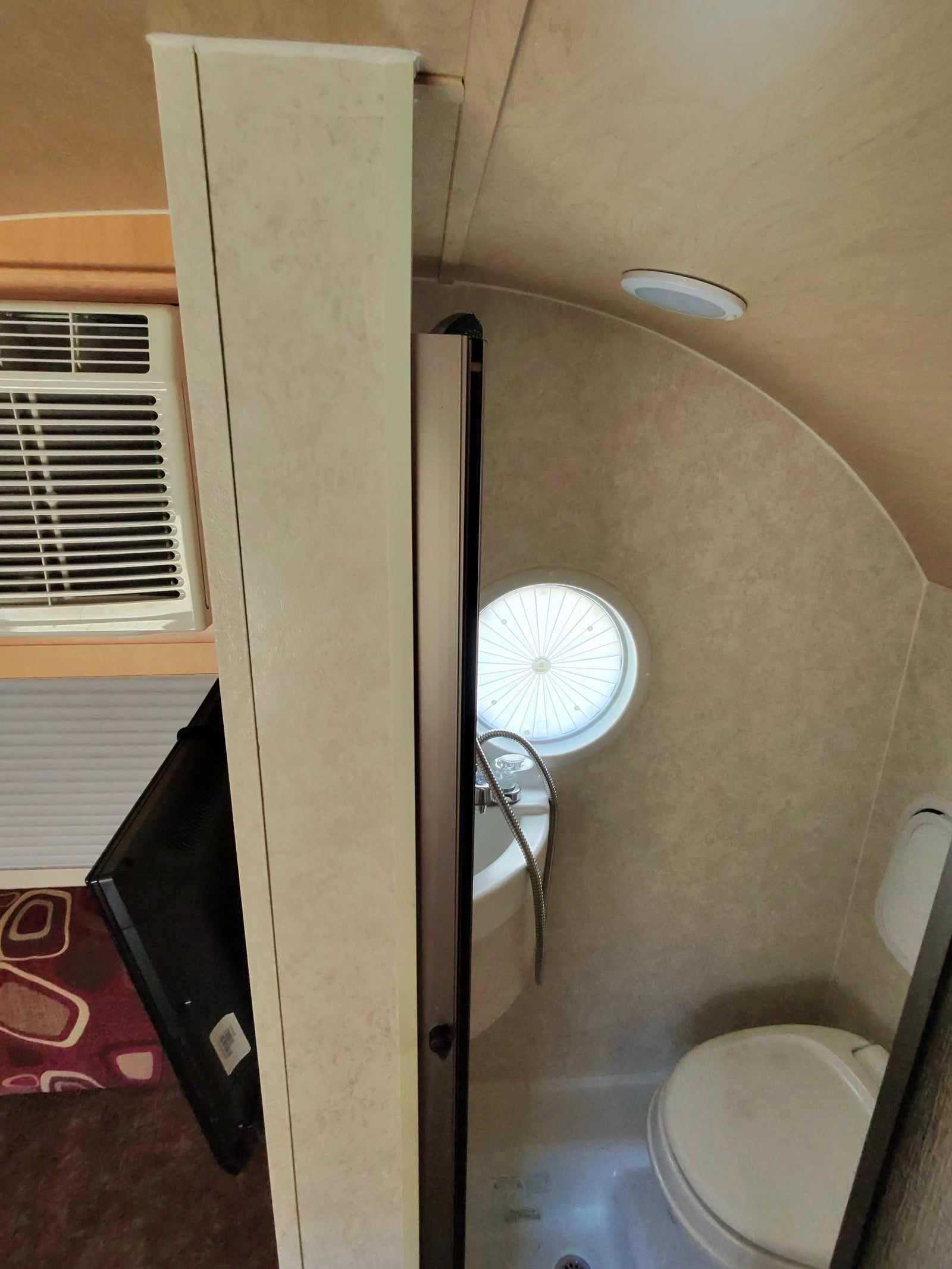 Compact rv rental interior shows a small wet bath with toilet, sink, circular window, and a wall-mounted AC unit.