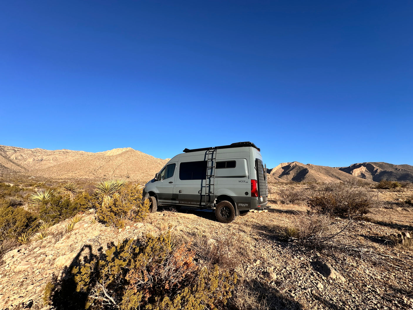 This grey Storyteller Overland 4x4 adventure van, ideal for an RV rental, is parked on a desert hill with arid mountains.
