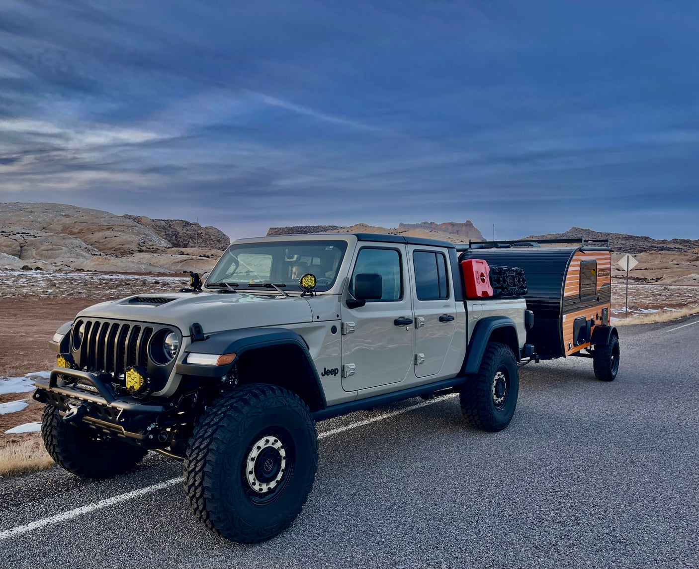 Light-colored Jeep Gladiator with an off-road RV rental trailer parked in a rugged desert landscape, ready for adventure.